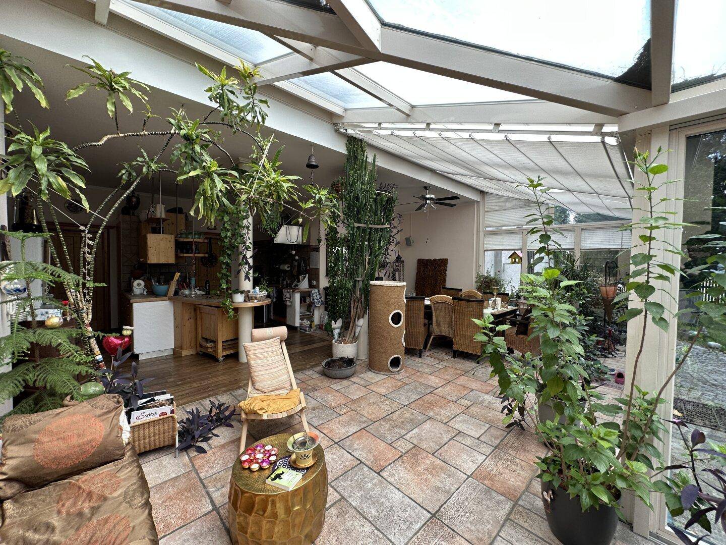 Heller Wintergarten mit vielen Pflanzen, Fliesenboden und gemütlichen Sitzgelegenheiten.