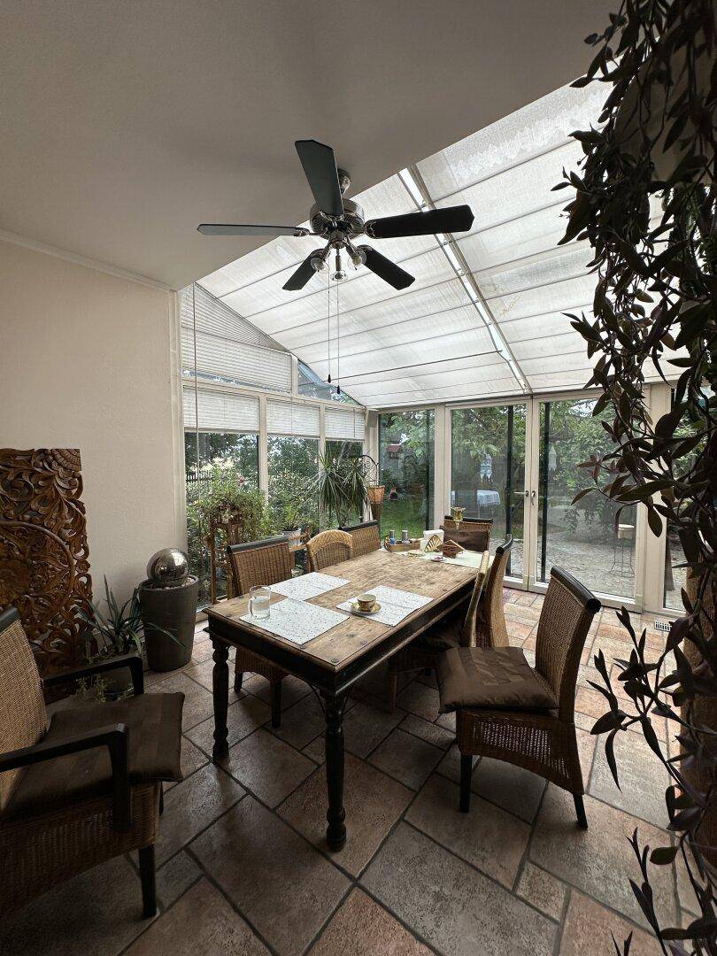 Essbereich im Wintergarten mit großem Holztisch, Korbstühlen und Deckenventilator.