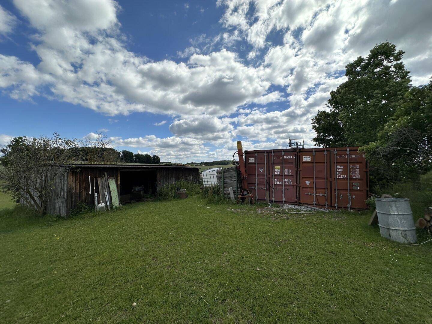 Altes Holzschuppen und Lagercontainer auf einer grünen Wiese unter blauem Himmel.