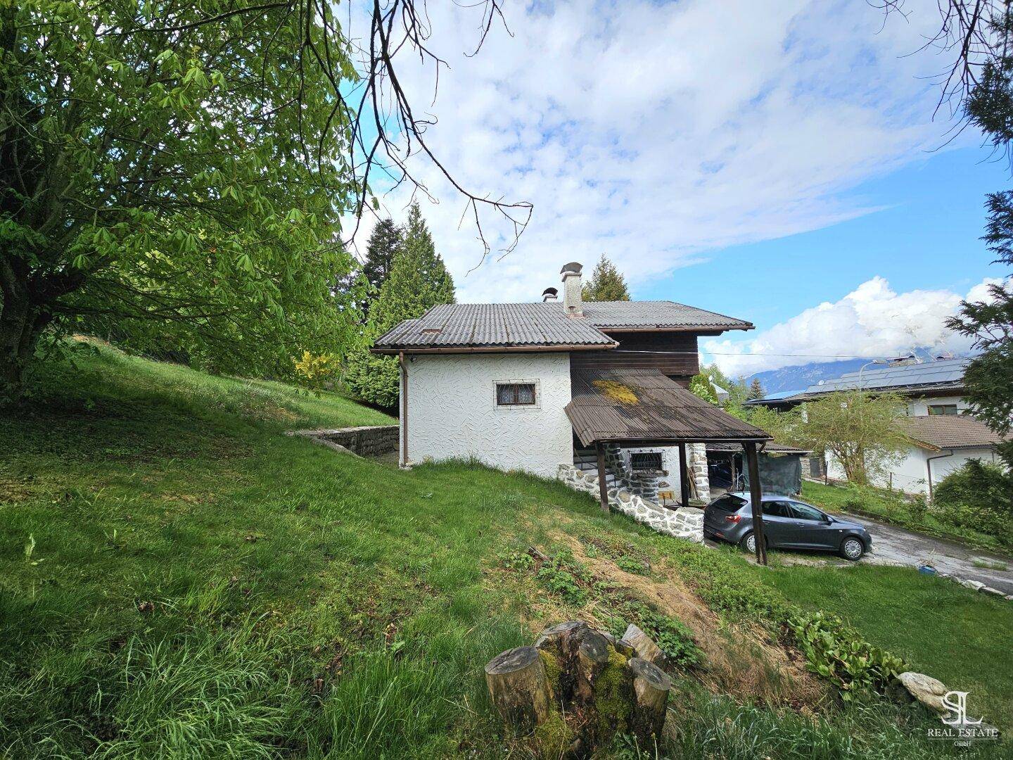 Seitenansicht des Hauses mit weißer Fassade, umgeben von einem grünen Hang und Bäumen.