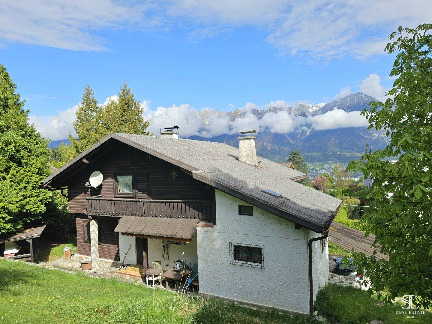 Haus mit Holzbalkon und weißer Fassade, eingebettet in eine grüne Umgebung mit Bergblick.