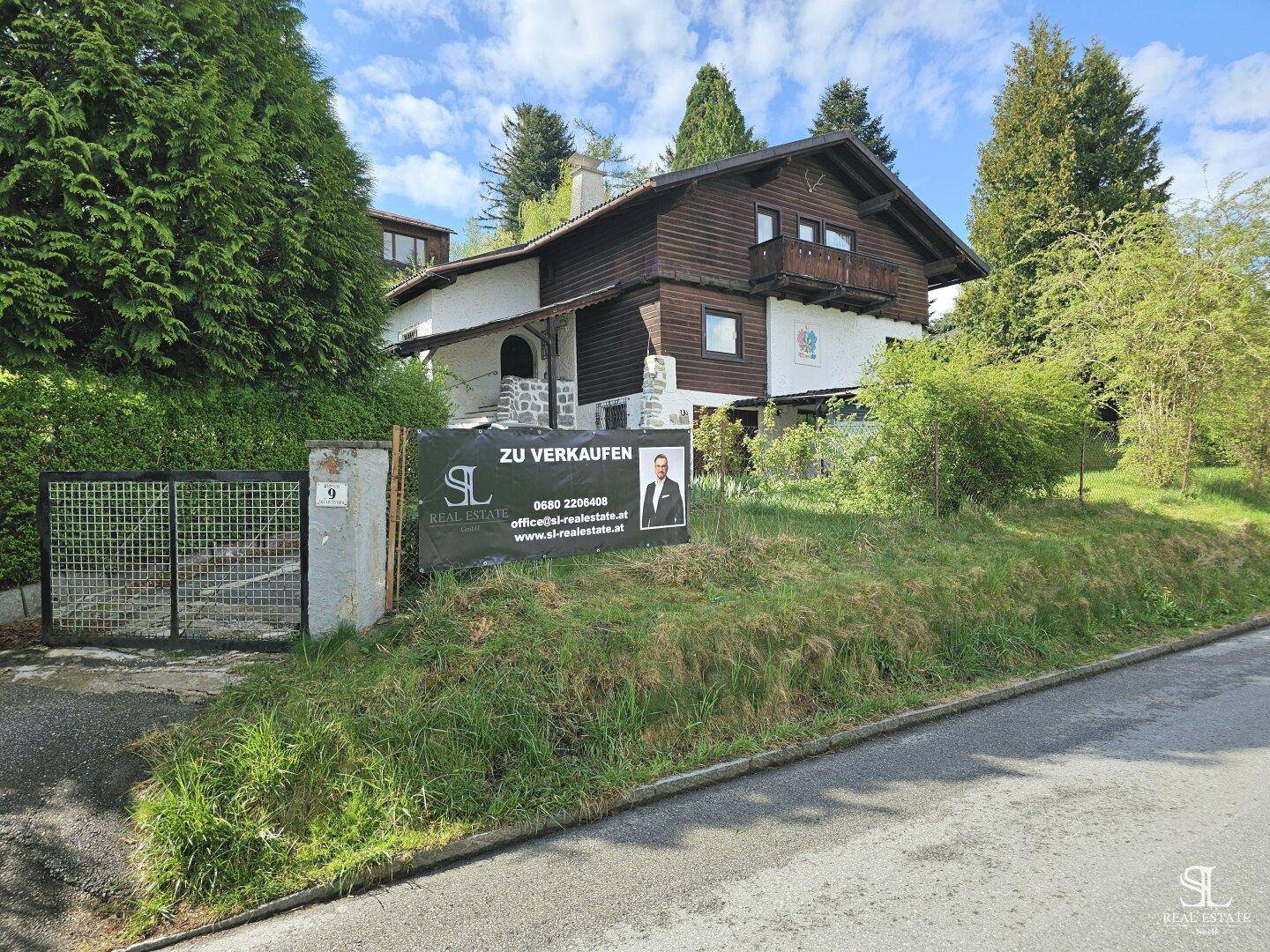 Hausansicht von der Straße mit einem "Zu Verkaufen"-Schild und grüner Umgebung.