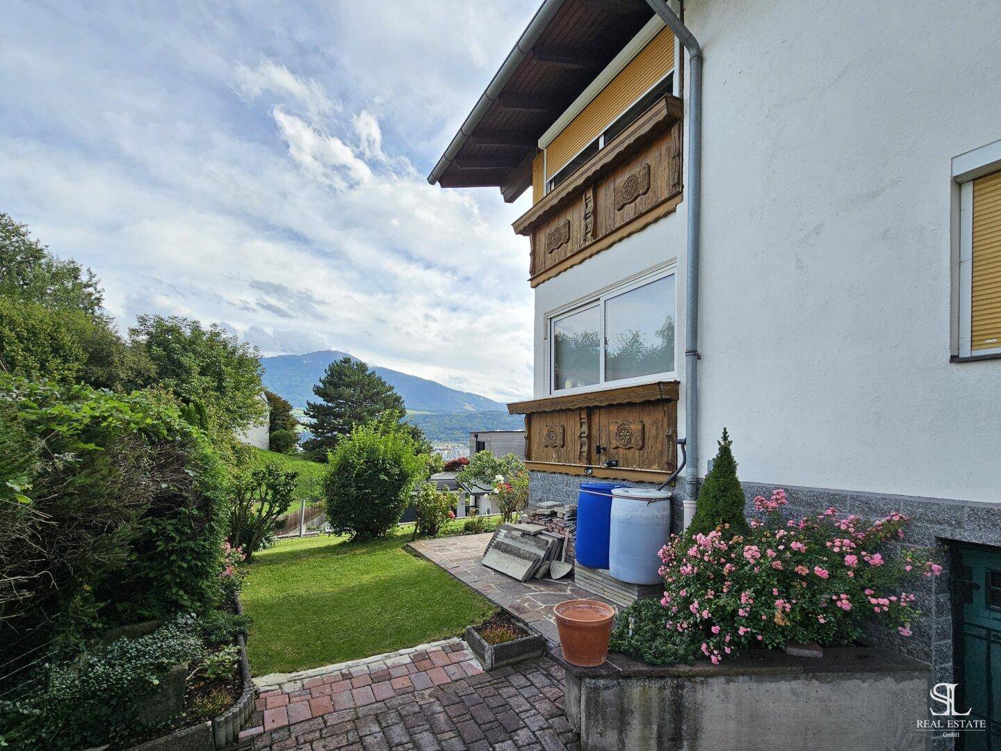 Seitlicher Zugang zum Haus mit gepflastertem Weg, Blumenbeeten und Blick auf die Berge.