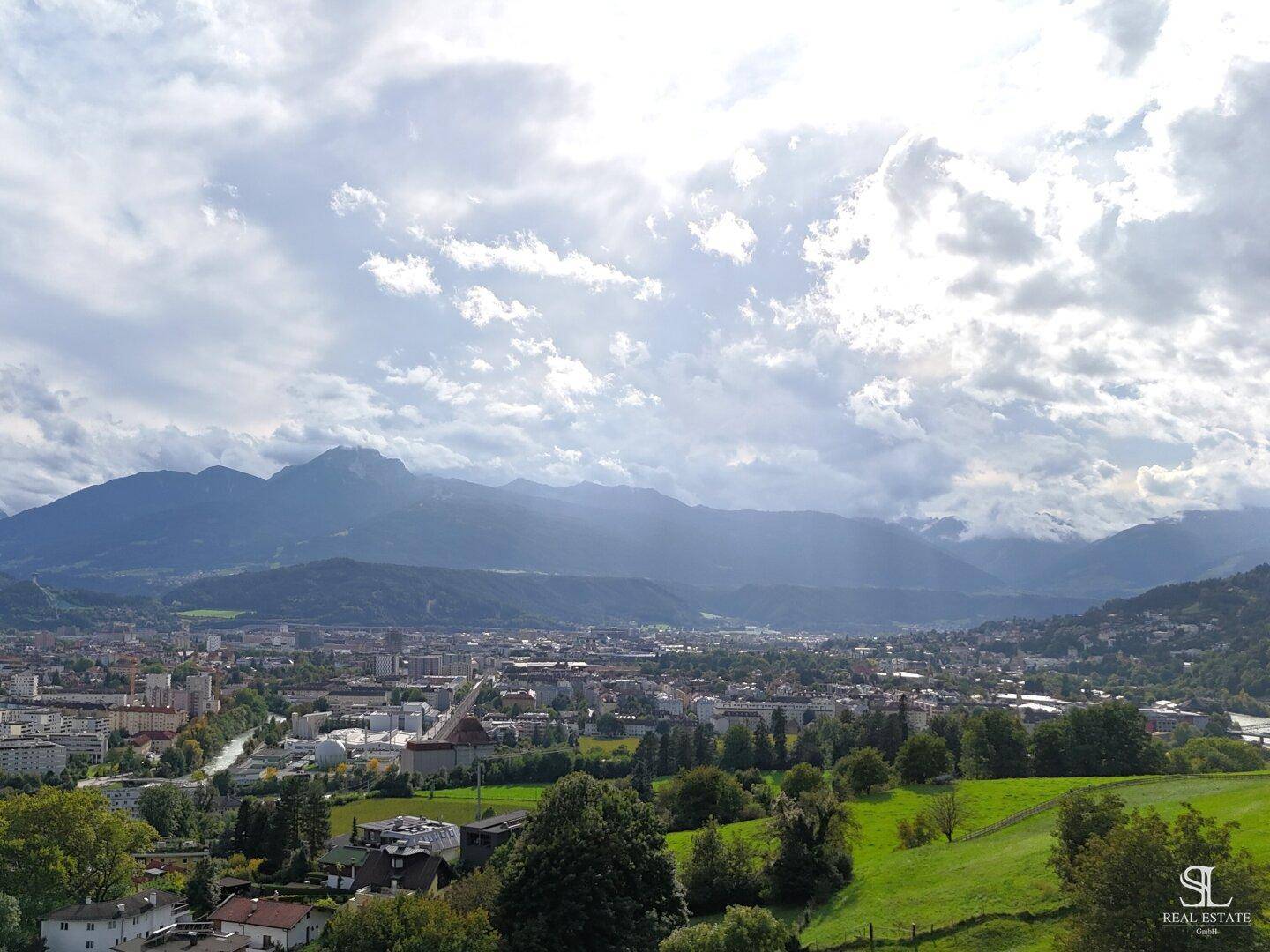 Panoramablick auf die Stadt und die umliegenden Berge unter einem dramatischen Wolkenhimmel.