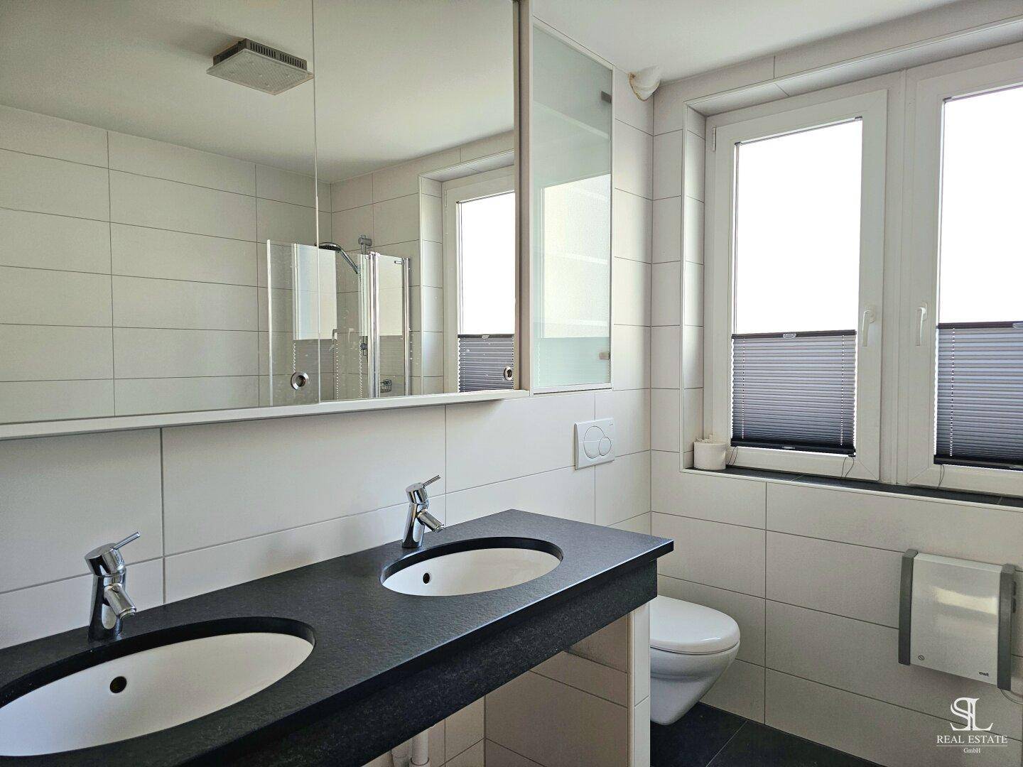 Helles Badezimmer mit Doppelwaschbecken, großem Spiegel, Toilette und Fenster für Tageslicht.
