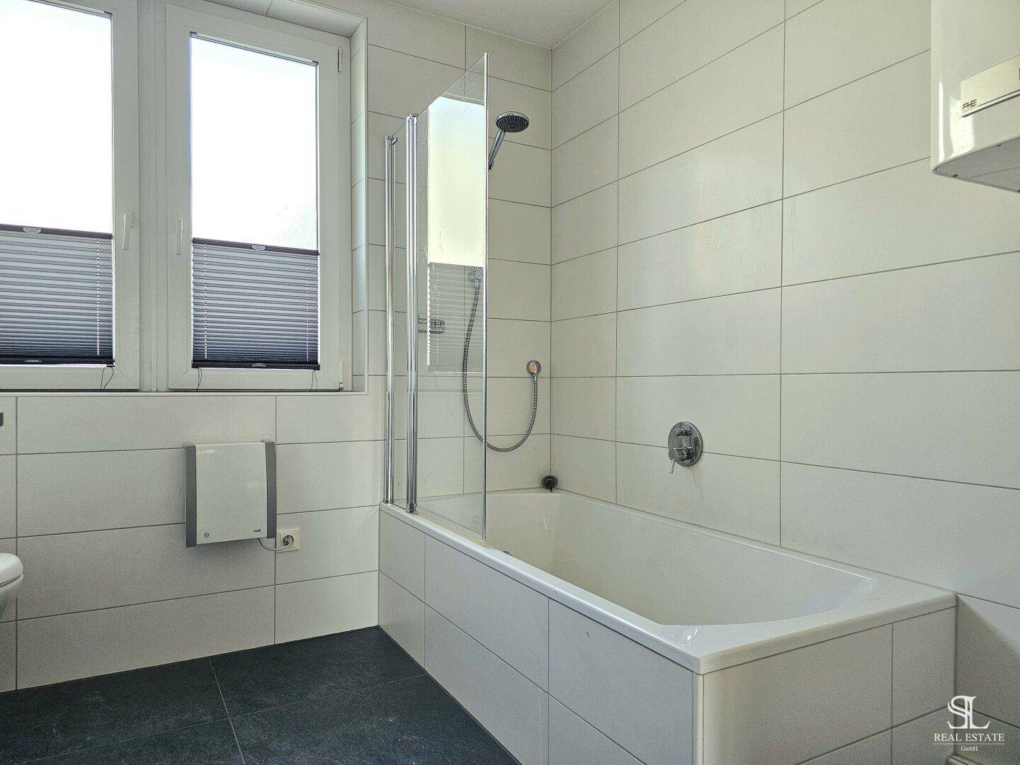 Detailansicht der Badewanne mit Duschabtrennung, umgeben von weißen Fliesen und dunklem Boden.