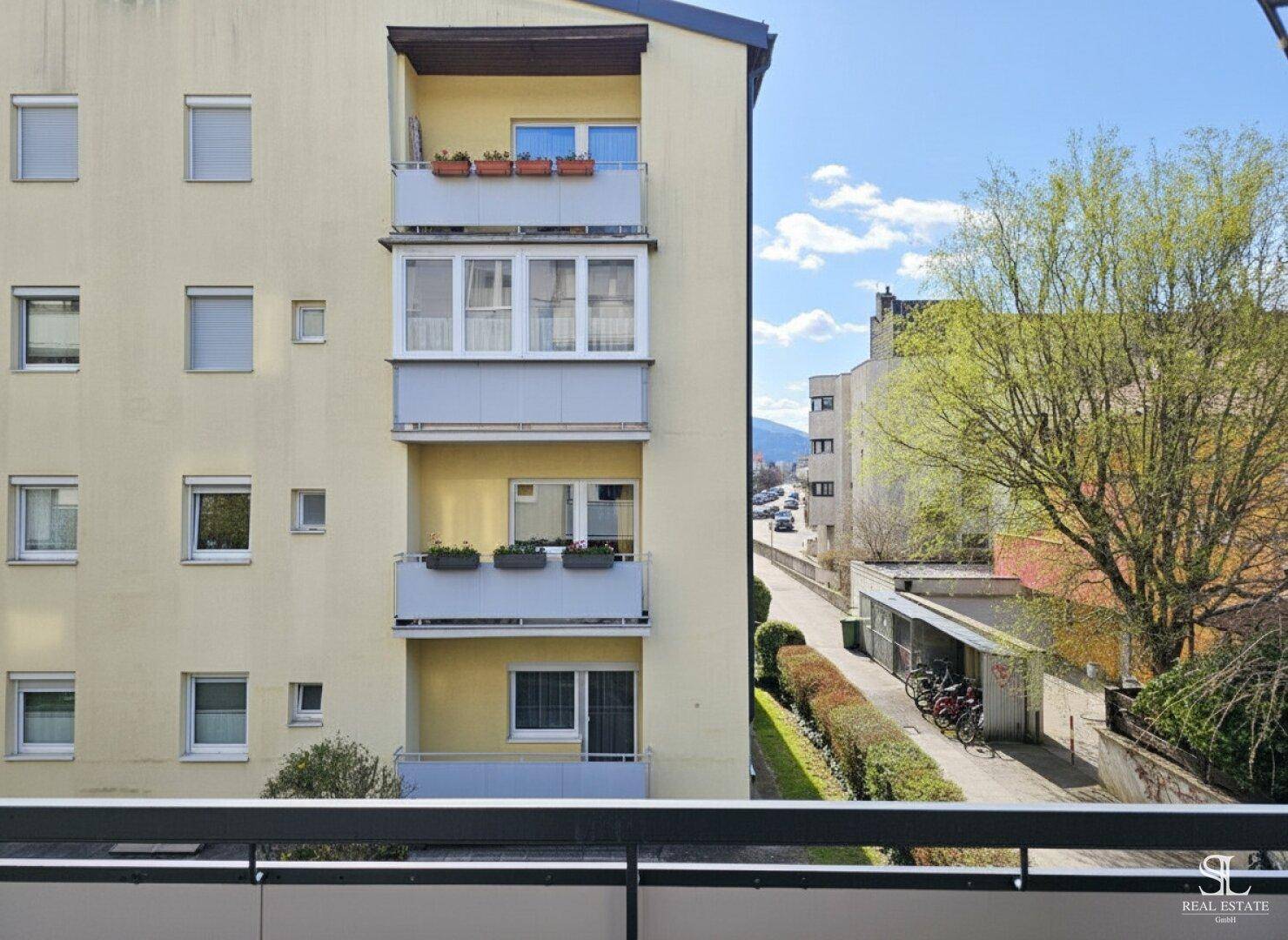 Blick vom Balkon auf eine Wohnhausfassade und die grüne Umgebung mit Bäumen.