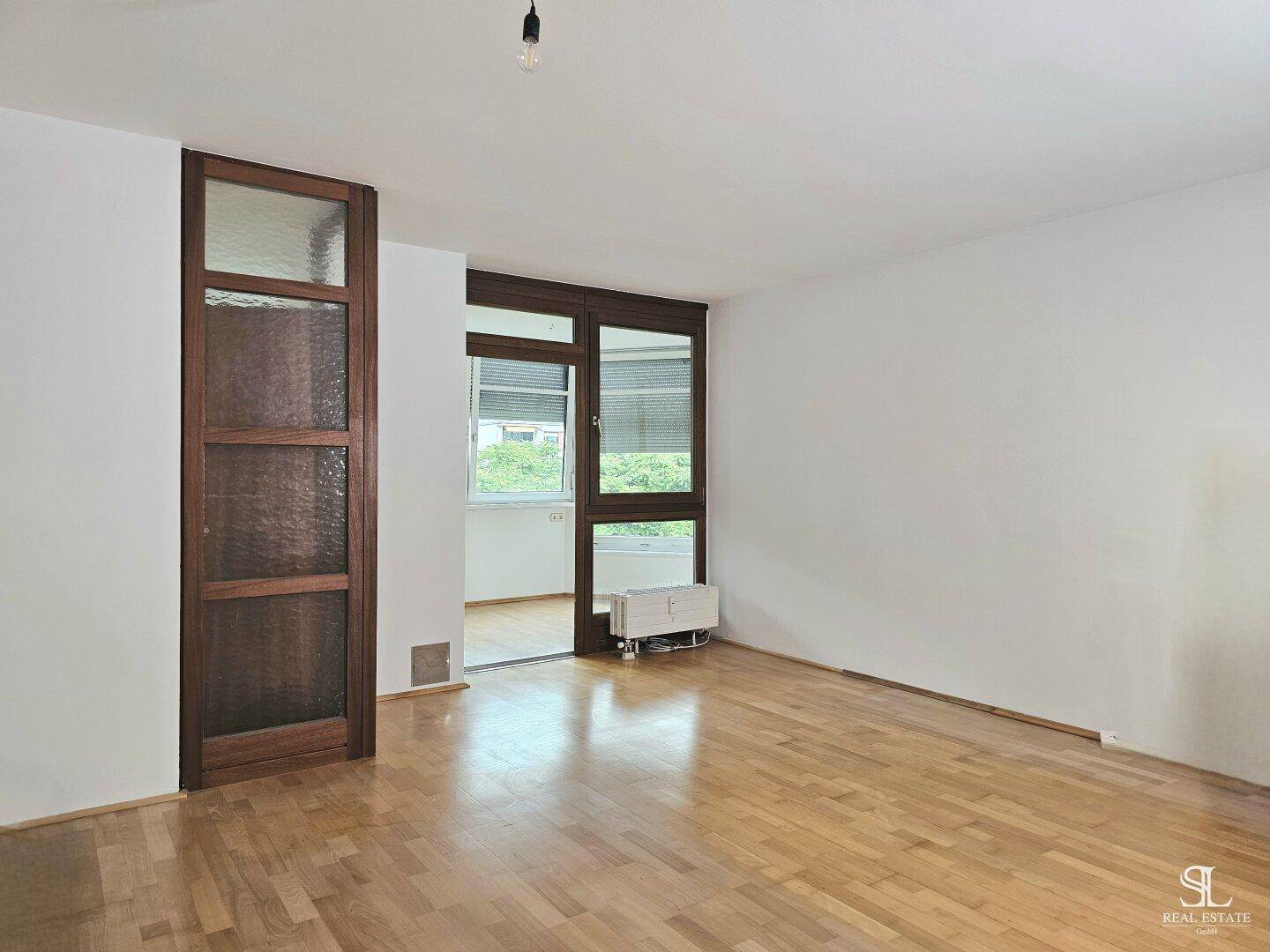 Leerer Raum mit hellem Holzboden und Zugang zu einem weiteren Bereich durch eine große Fensterfront.
