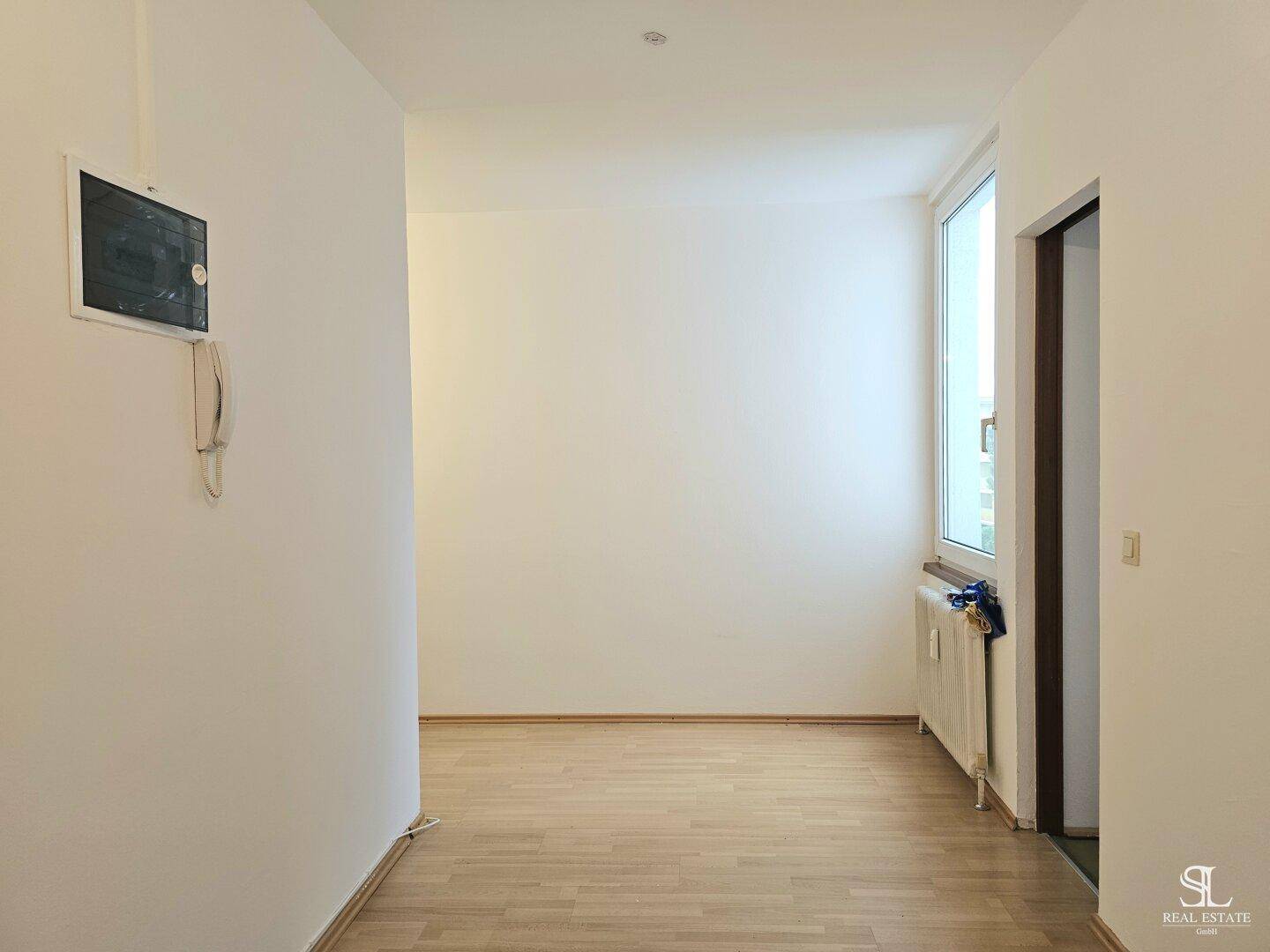 Leeres Zimmer mit Holzboden, Fenster und Heizkörper, ausgestattet mit einer Gegensprechanlage.