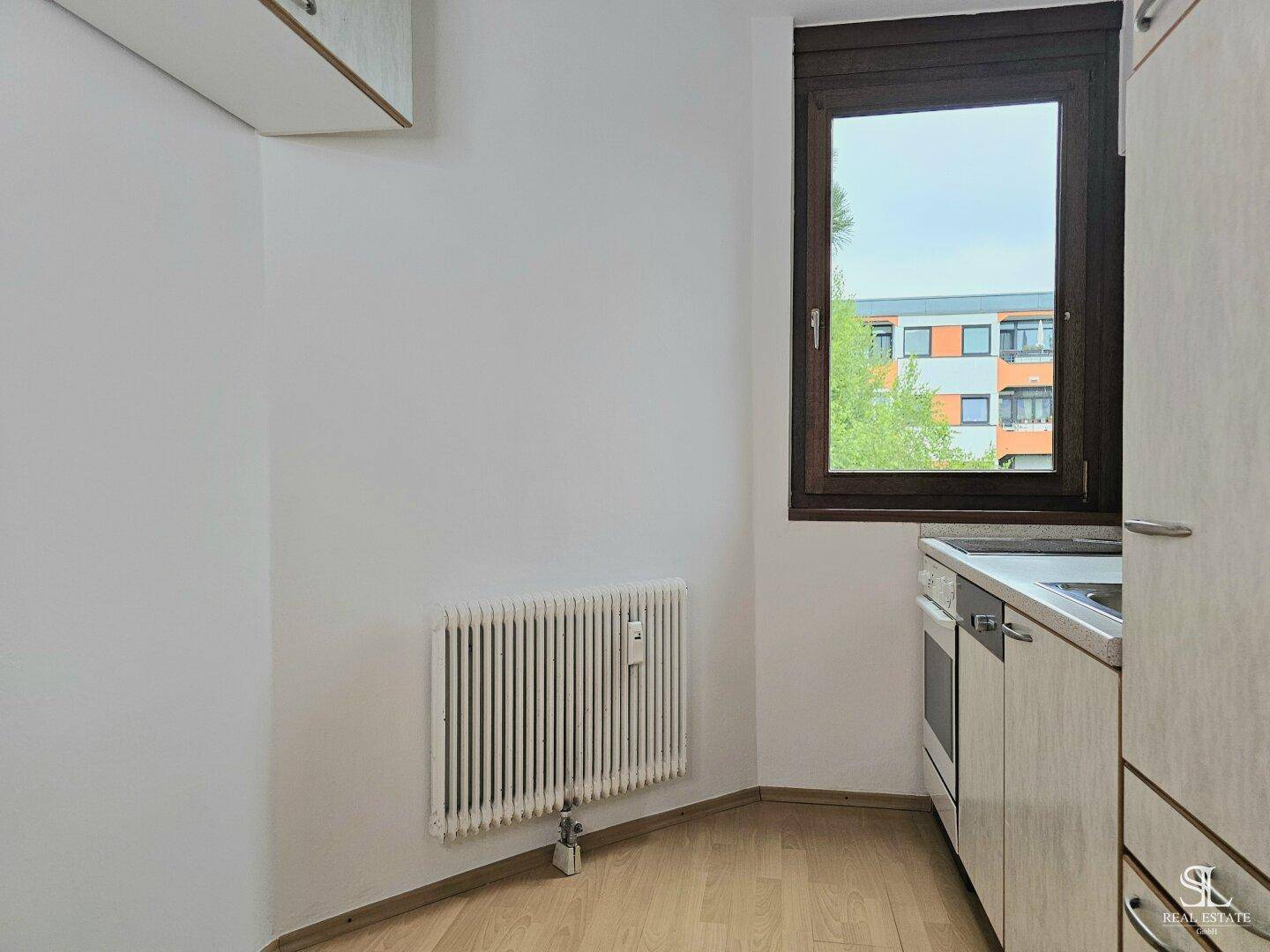 Kleine Küche mit Einbauschränken, Spüle, Kochfeld und Fenster, das Tageslicht hereinlässt.