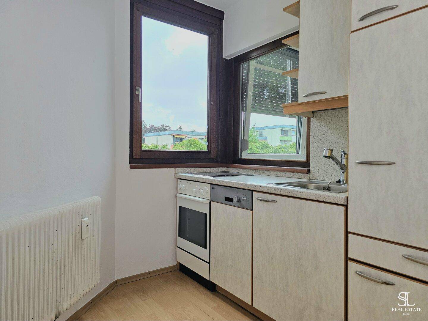 Kompakte Küche mit hellen Schränken, Spüle, Kochfeld und Fenster, das den Raum erhellt.
