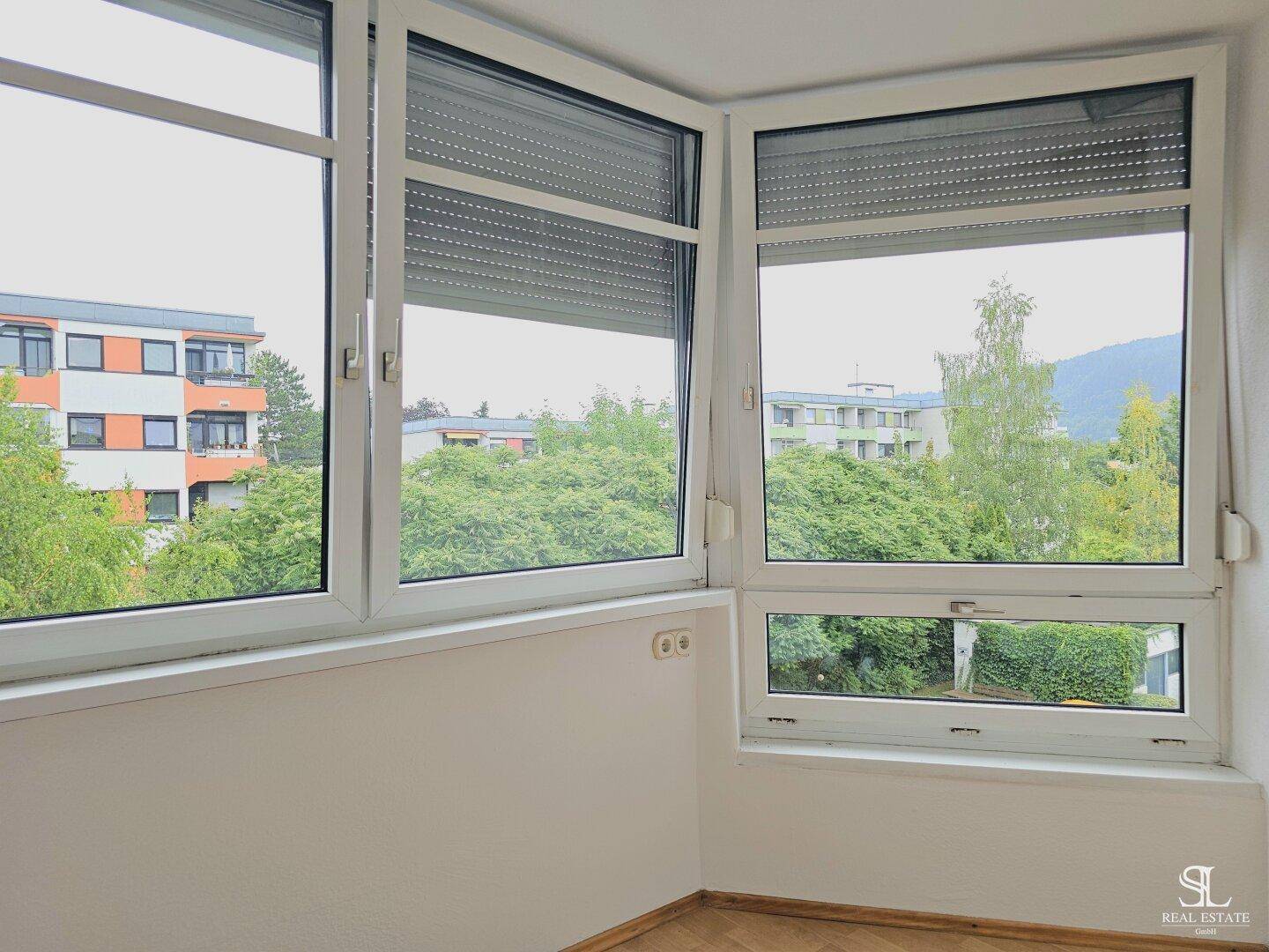 Zimmer mit mehreren Fenstern und Rollläden, die einen Ausblick auf Bäume und Gebäude bieten.