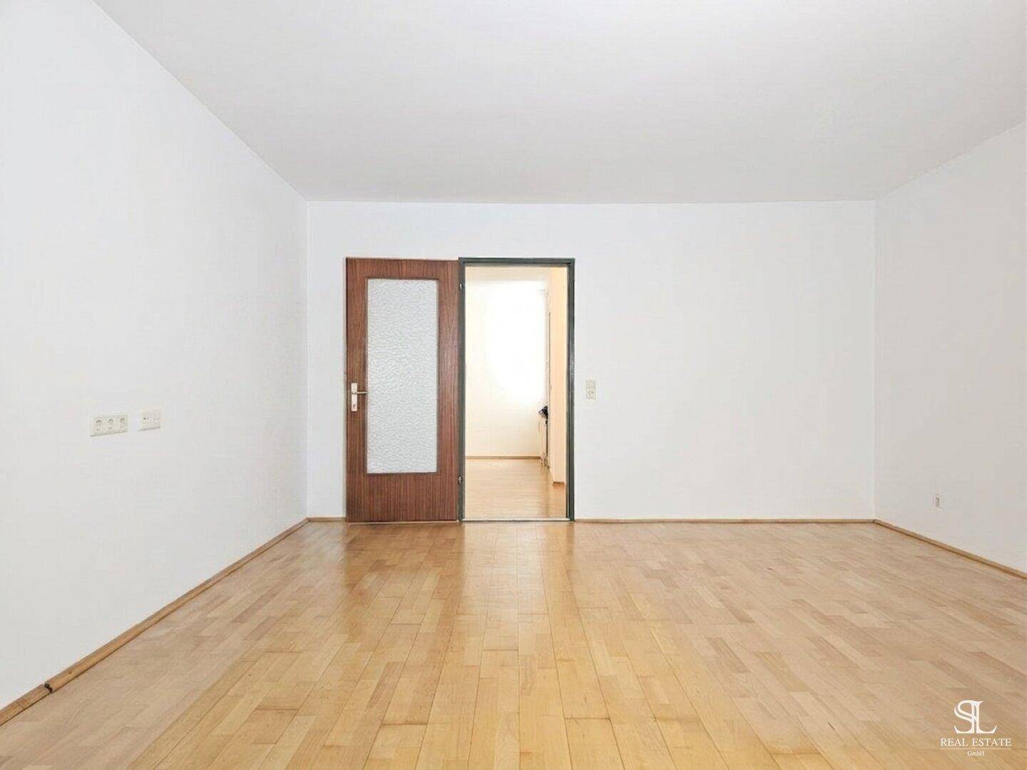 Heller, leerer Raum mit Holzboden und einer Tür, die zu einem angrenzenden Flur führt.