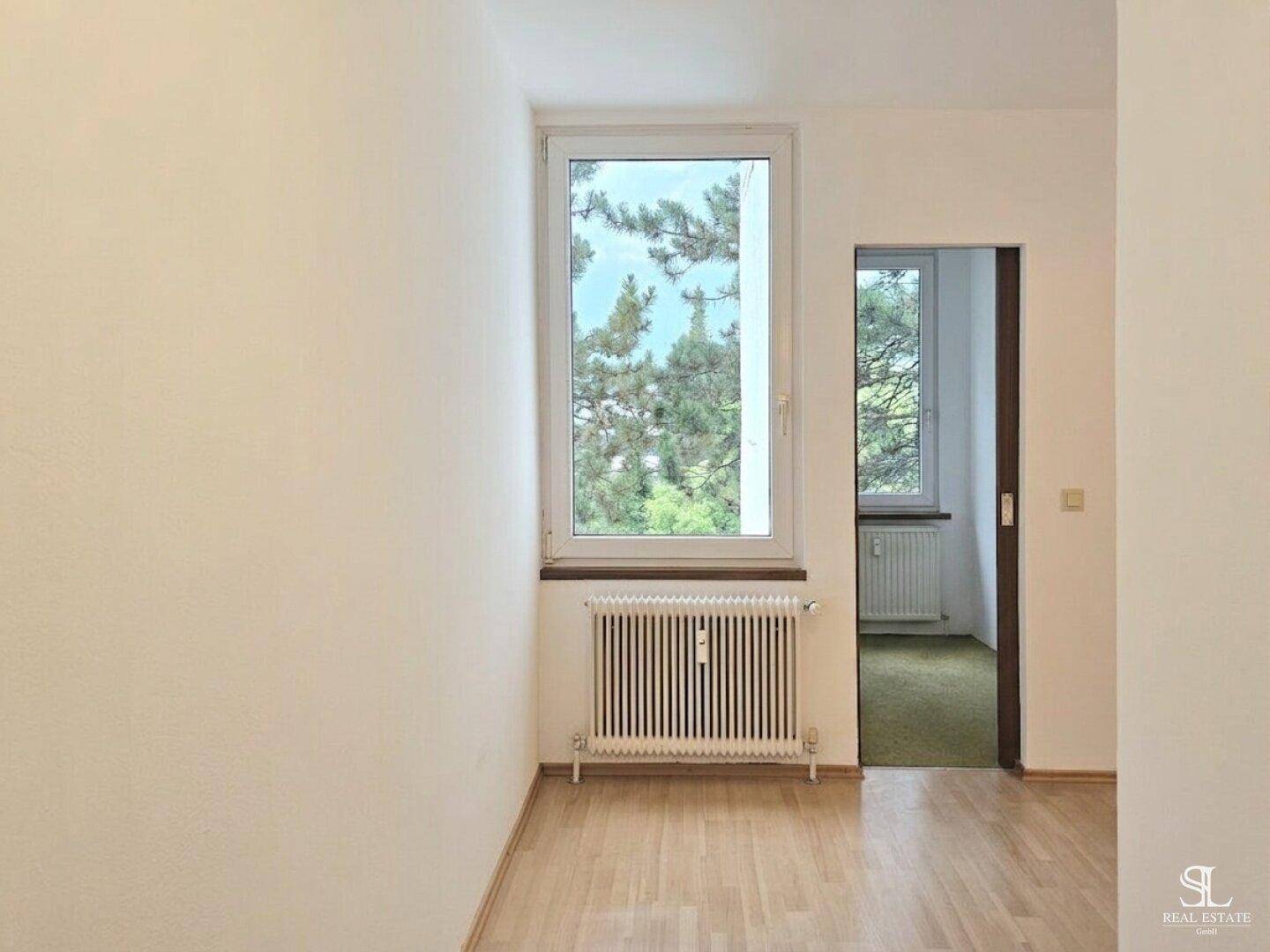 Helles Zimmer mit Holzboden, Fenster und Heizkörper, bietet Blick ins Grüne.
