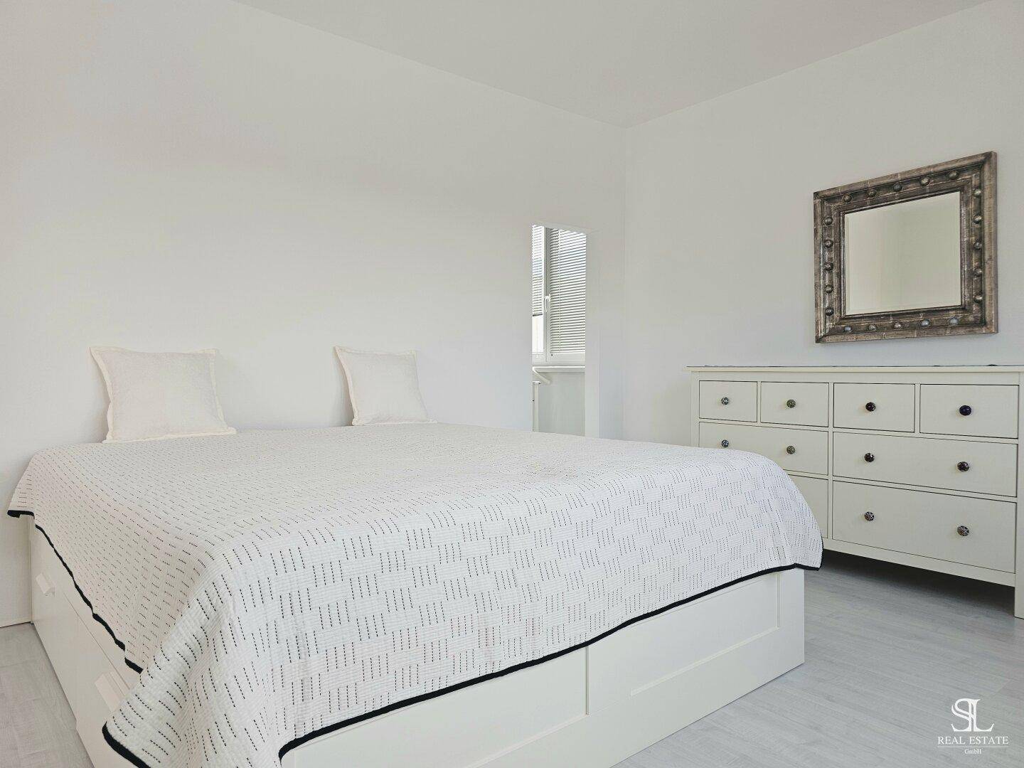 Schlafzimmer mit weißem Bett und Kommode, minimalistisch eingerichtet.