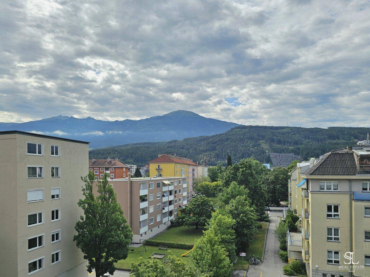 Panoramablick über Wohngebäude, grüne Flächen und majestätische Berge unter bewölktem Himmel.