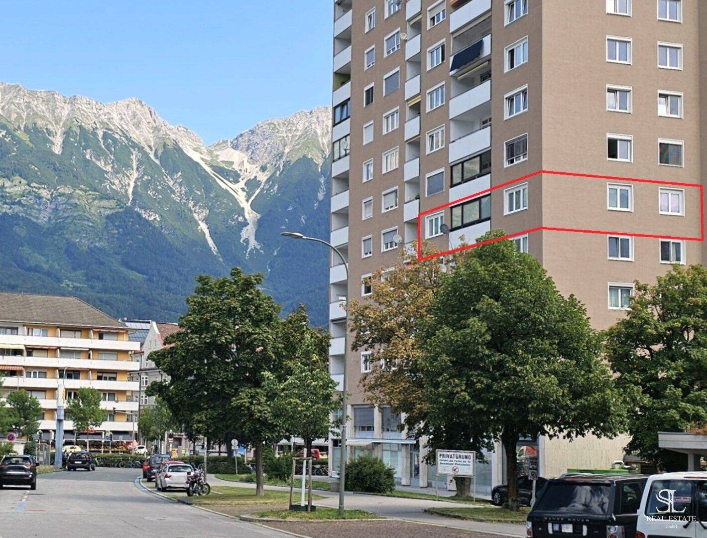 Hohes Wohngebäude mit Balkonen an einer Straße, umgeben von beeindruckenden Bergen.