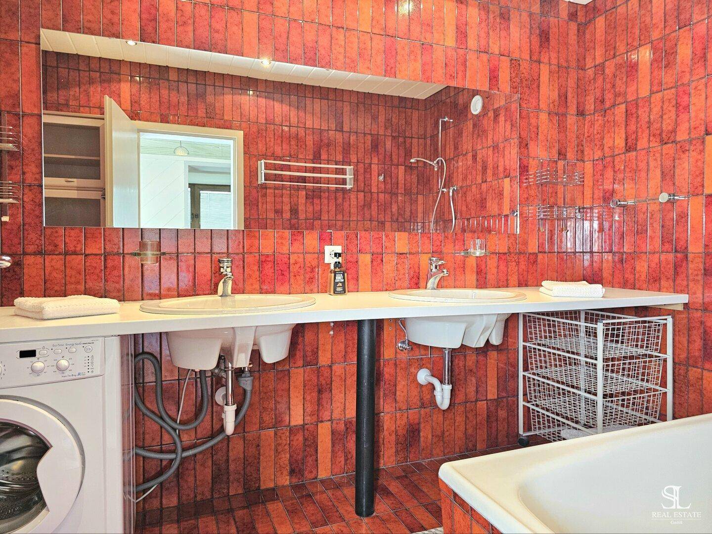 Badezimmer mit orange-braunen Fliesen, Doppelwaschbecken, großem Spiegel und Waschmaschine.