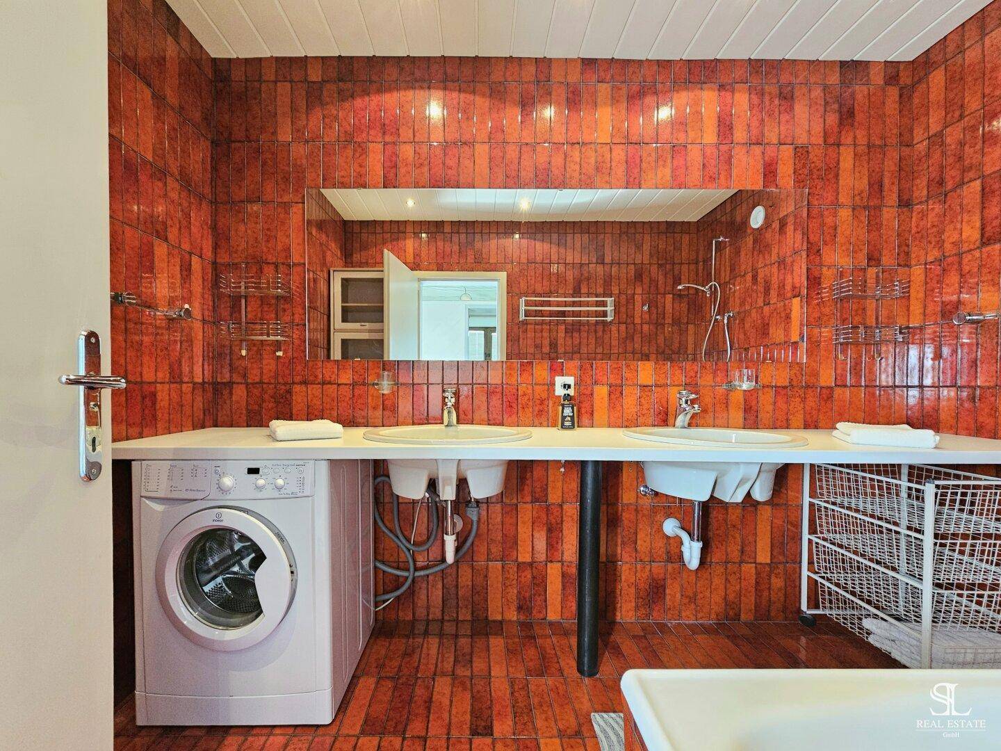 Geräumiges Badezimmer mit orange-braunen Fliesen, Doppelwaschbecken, großem Spiegel und Waschmaschine.