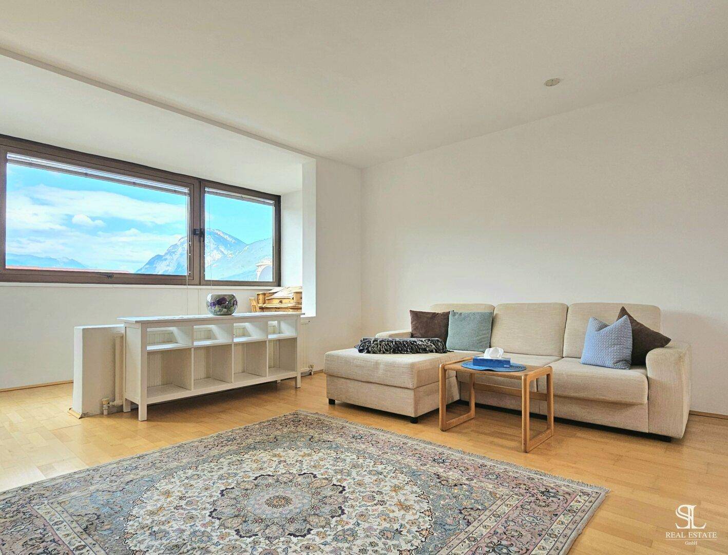 Gemütliches Wohnzimmer mit hellem Parkettboden, beiger Couch und weitem Blick auf die Berge.