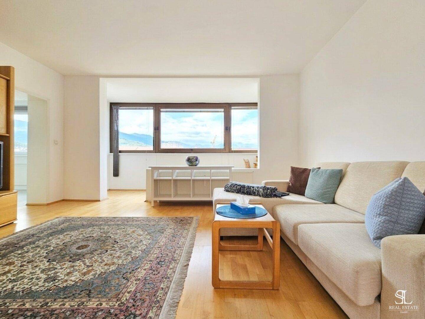 Geräumiges Wohnzimmer mit beigem Sofa, Teppich und Blick auf die umliegende Berglandschaft.