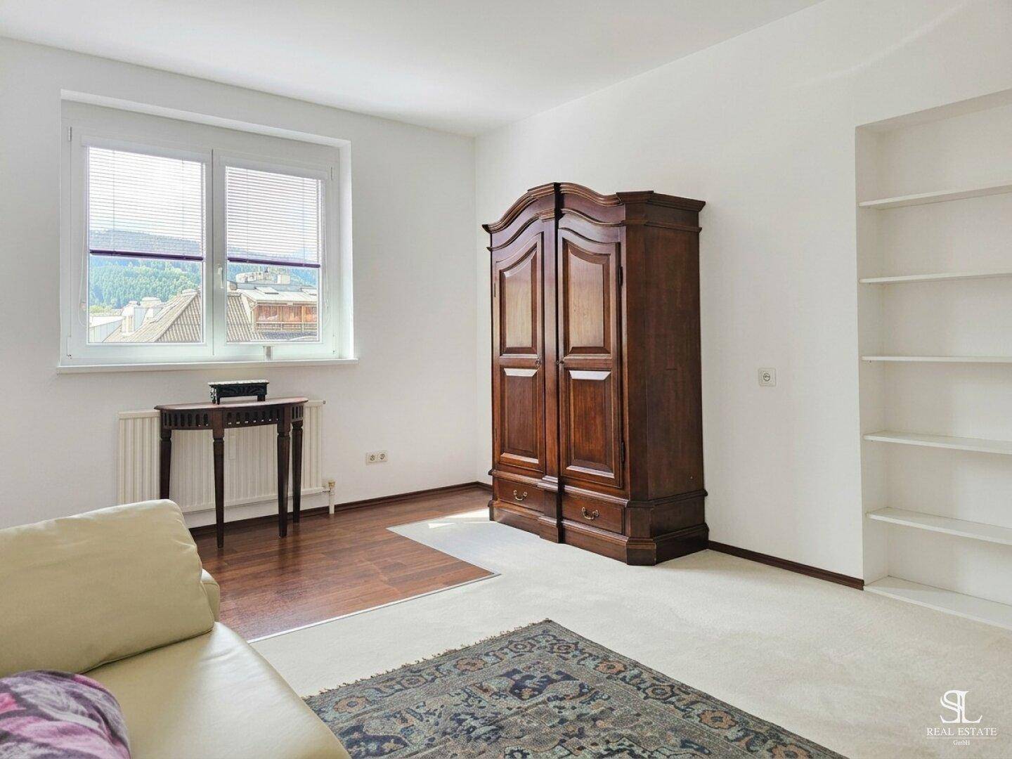 Heller Raum mit großem Holzschrank, kleinem Tisch am Fenster und Blick auf die Umgebung.