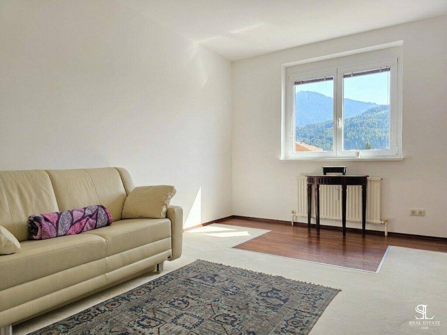 Helles Wohnzimmer mit beigem Sofa, Teppich und Fenster mit Bergblick, teilweise Holzboden.