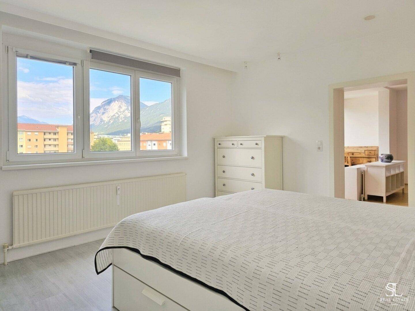 Helles Schlafzimmer mit weißem Bett, Kommode und weitem Blick auf die Berge.