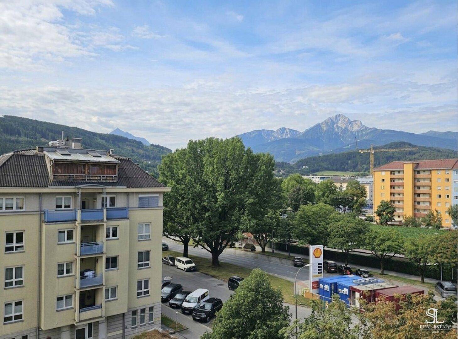 Weitläufiger Blick über die Stadt mit grünen Bäumen und majestätischen Bergen im Hintergrund.
