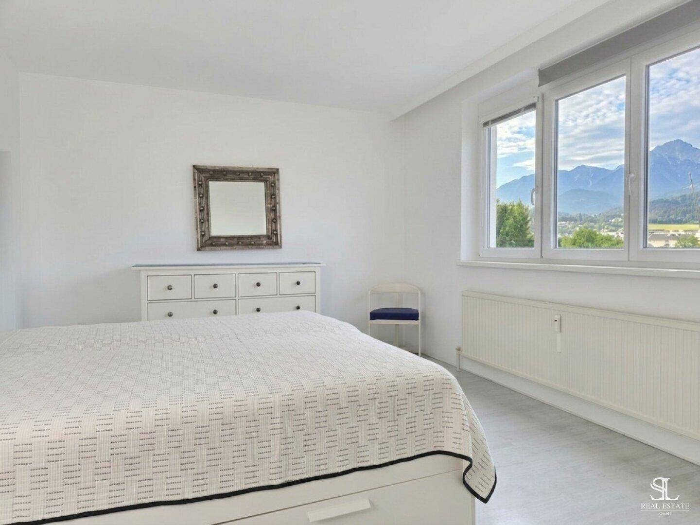 Geräumiges Schlafzimmer mit weißem Bett, Spiegel und Fenster mit Bergblick.