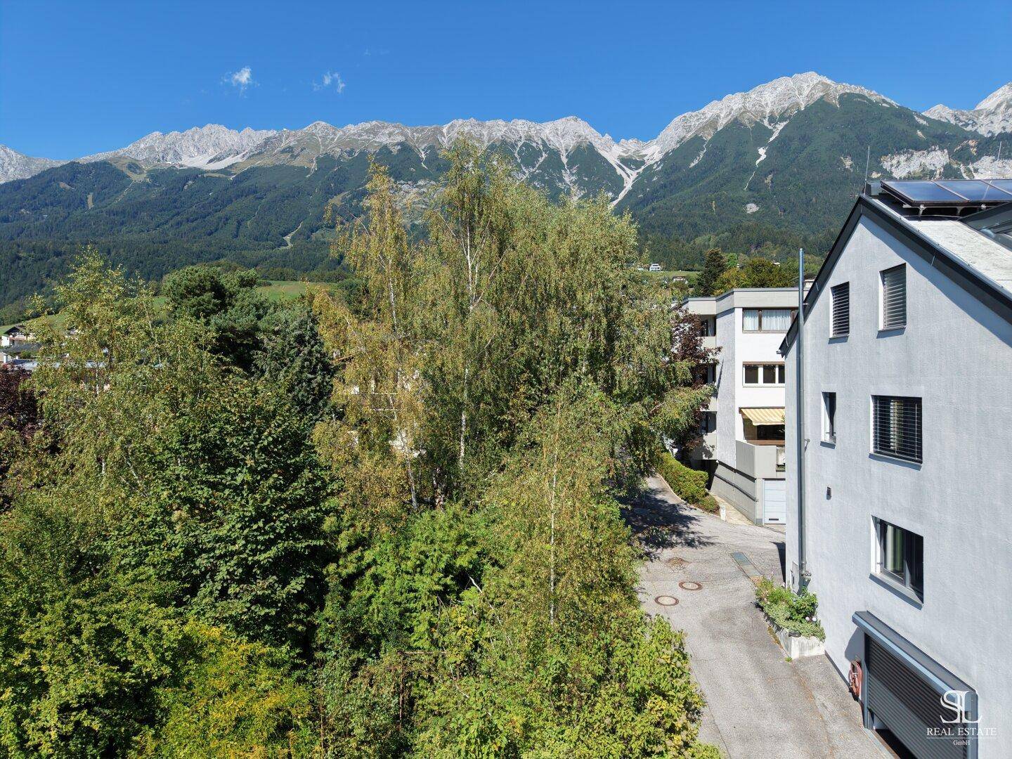 Blick auf eine ruhige Straße, umgeben von Bäumen und Gebäuden, mit majestätischen Bergen im Hintergrund.