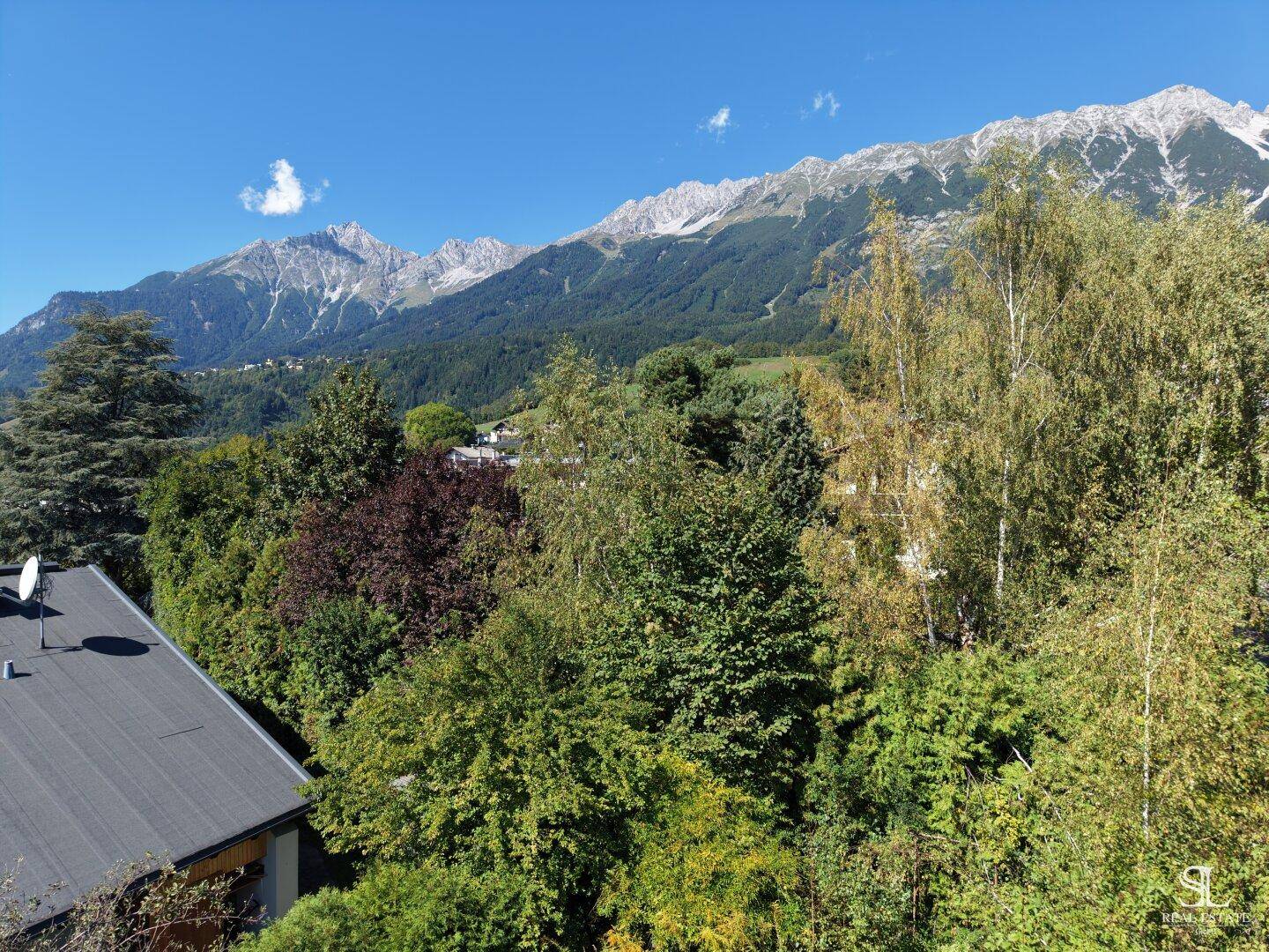 Grüne Umgebung mit Bäumen und Häusern, die sich vor der imposanten Bergkette erstreckt.