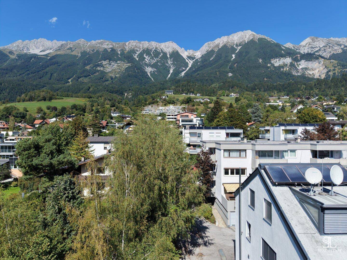 Weitblick über Wohngebäude und grüne Flächen mit den Alpen im Hintergrund.