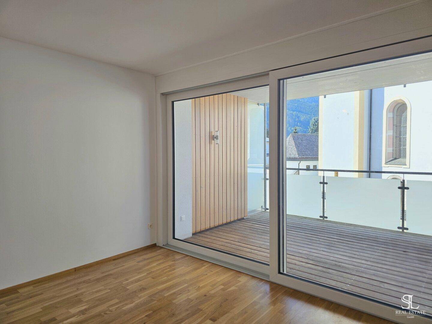 Heller Raum mit Holzboden und großer Schiebetür, die Zugang zu einem Balkon bietet.