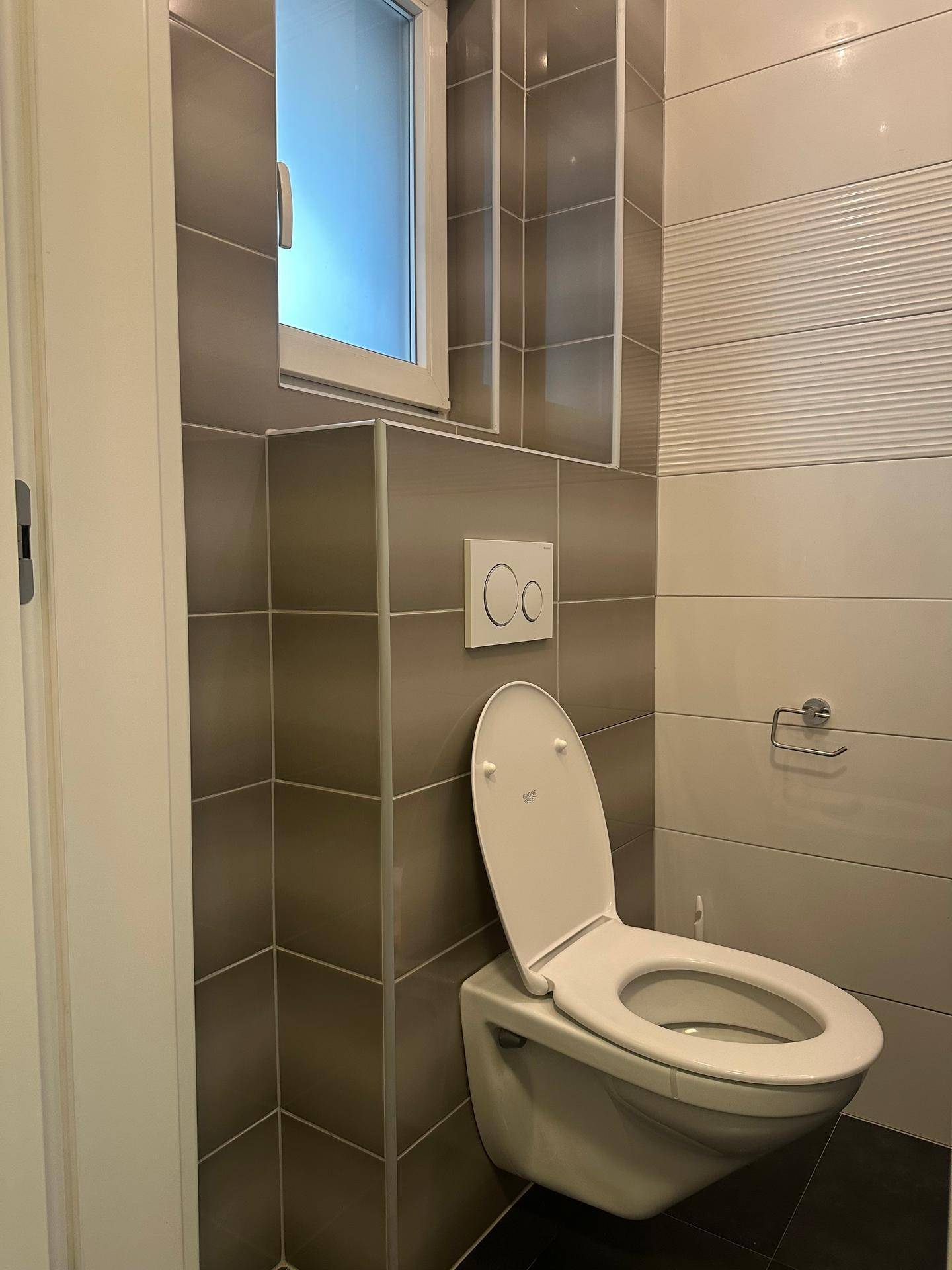 Helles Gäste-WC mit wandhängendem WC, Fenster und modernen Fliesen in Grau und Weiß.