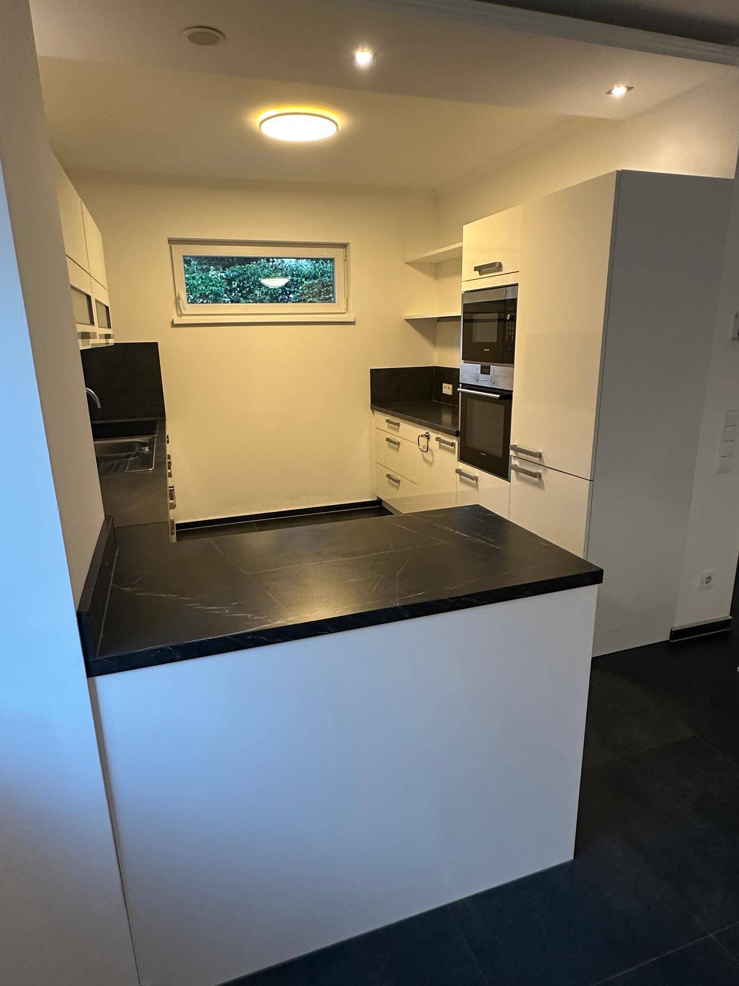 Moderne Küche mit weißen Hochglanzfronten, dunkler Arbeitsplatte und Fenster.