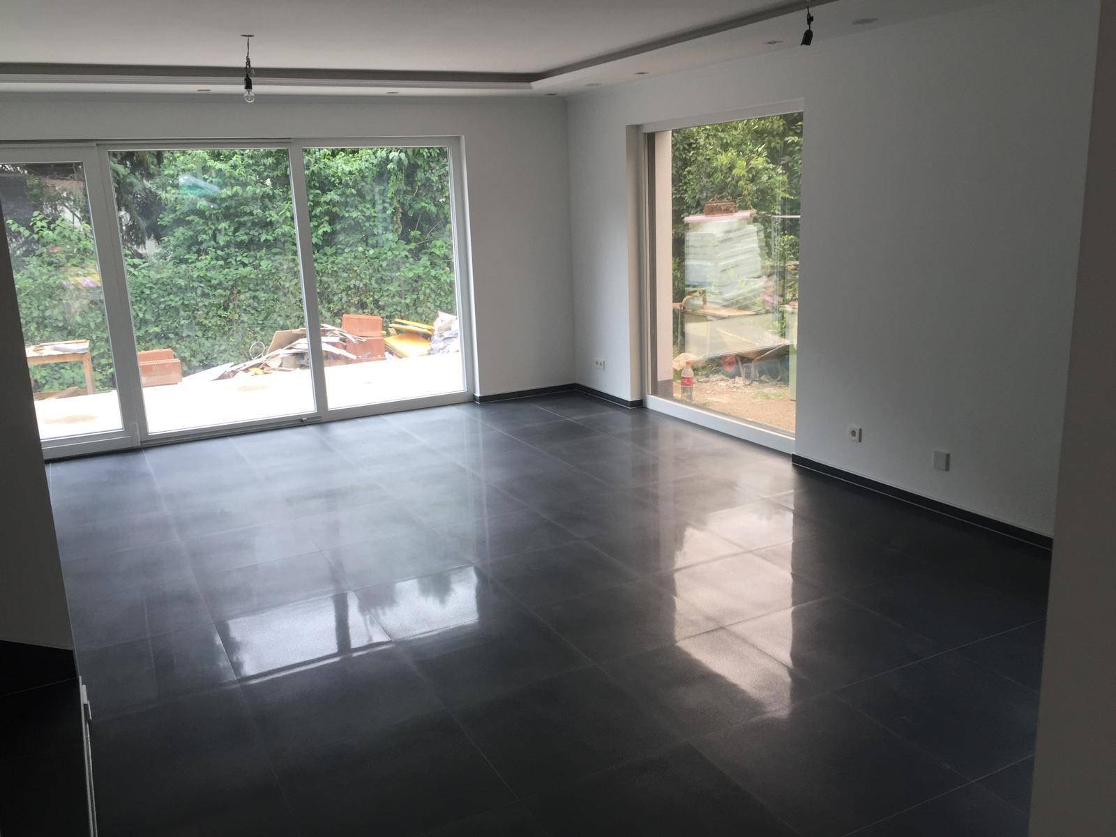 Geräumiges Wohnzimmer mit dunklem Fliesenboden und großen Fensterfronten zum Garten.