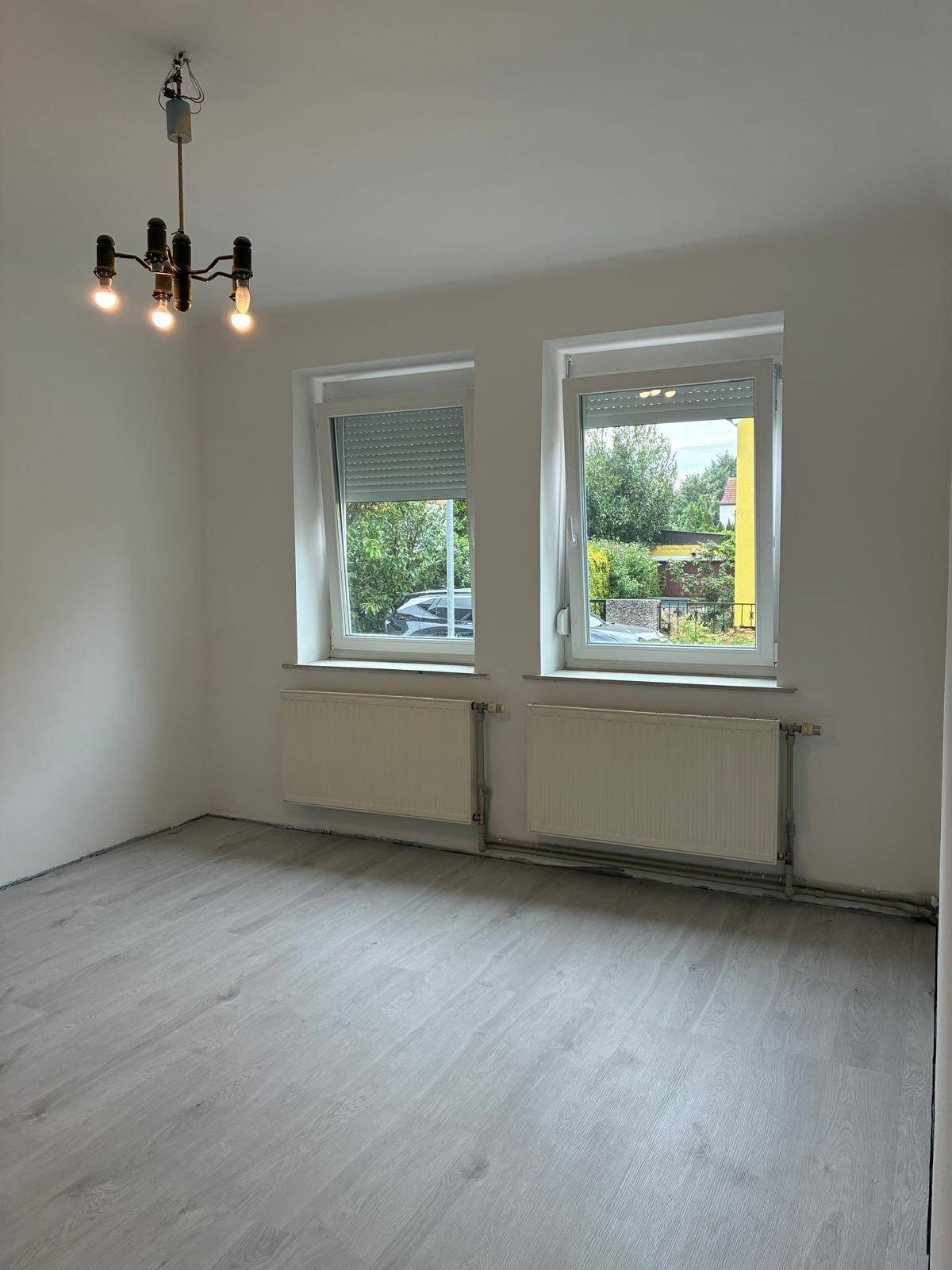 Leeres Wohnzimmer mit hellem Laminatboden, zwei Fenstern mit Rollläden und einem Deckenleuchter.