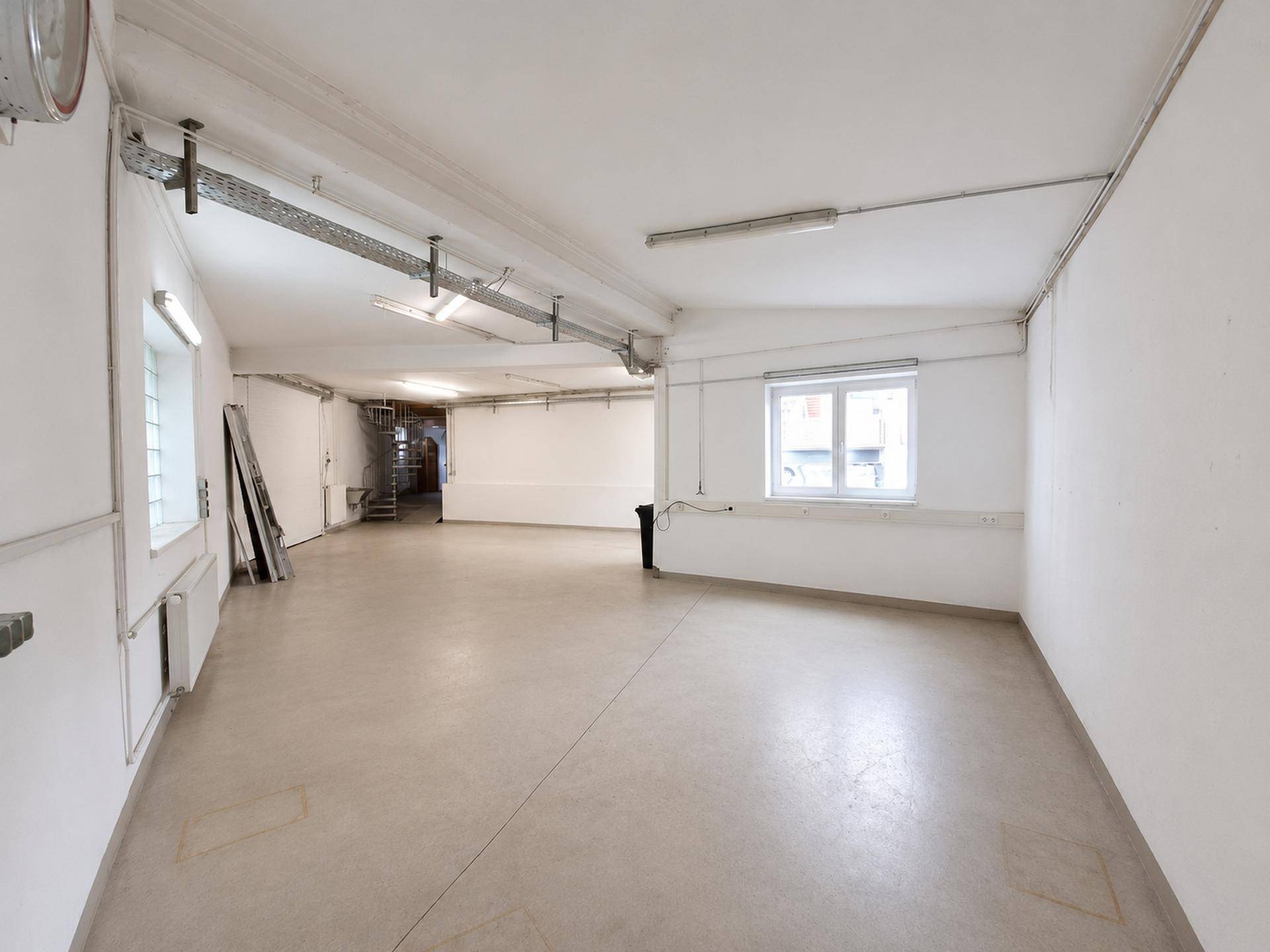 Großer, heller Gewerberaum mit Betonboden, Fenstern und Zugang zu einem Treppenhaus im Hintergrund.