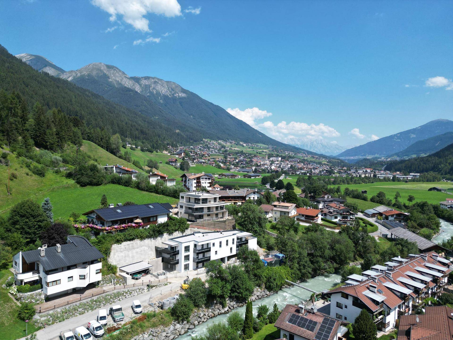 Weitläufige Luftaufnahme eines Tals mit Siedlung, Fluss und beeindruckender Bergkulisse unter blauem Himmel.