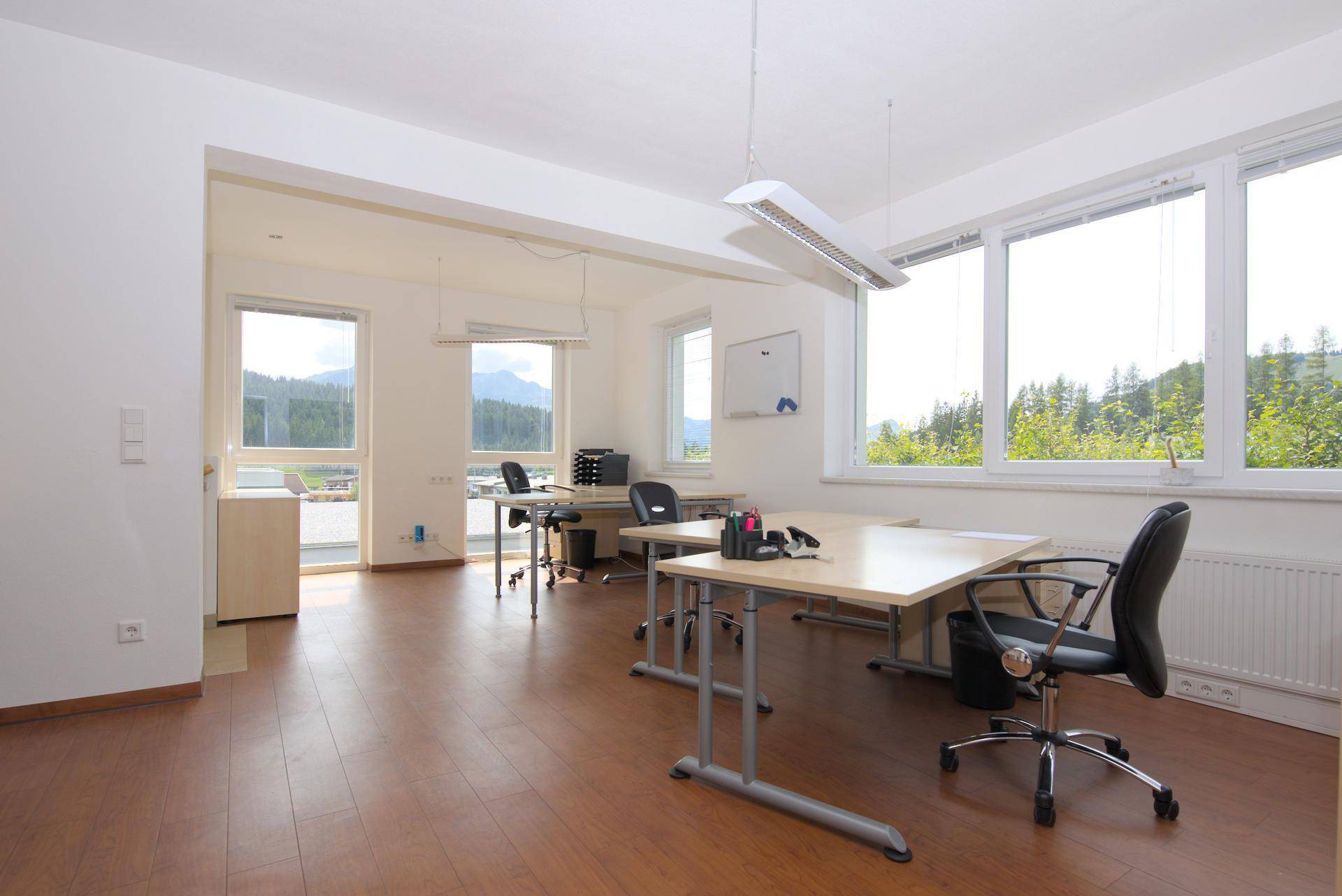 Helles Büro mit mehreren Arbeitsplätzen, großen Fenstern und Blick auf die umliegende Natur und Berge.