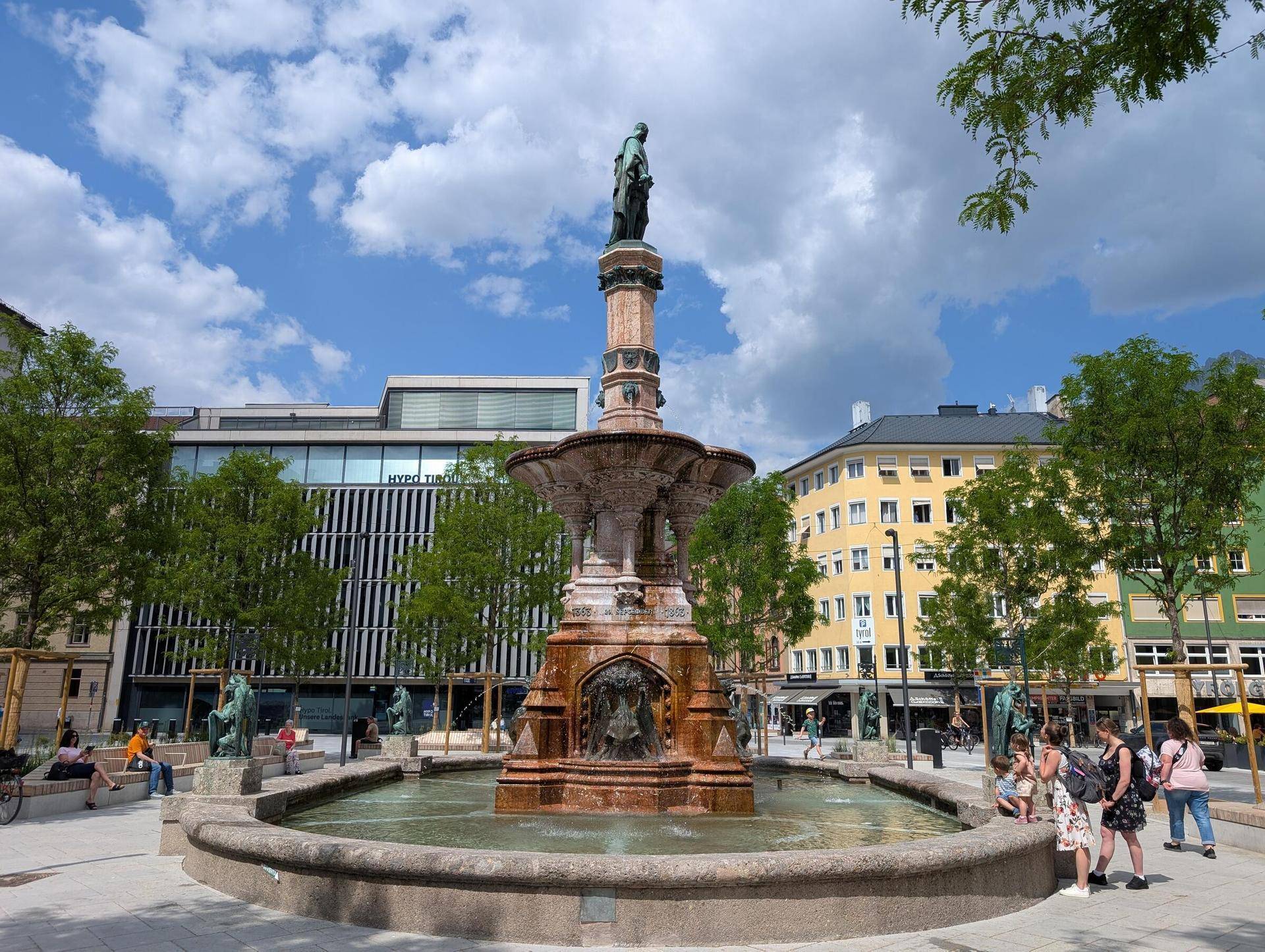 Historischer Brunnen auf einem belebten Stadtplatz, umgeben von Gebäuden und Bäumen unter blauem Himmel.