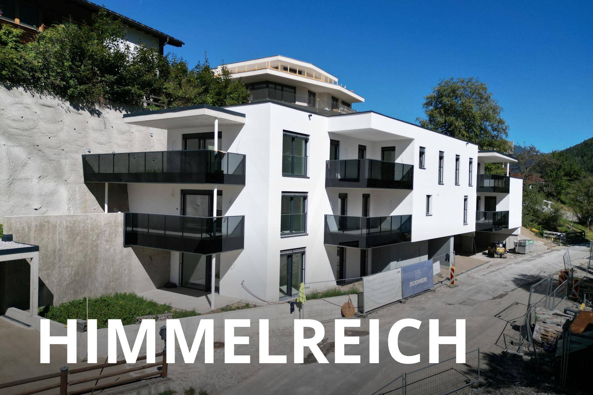 Modernes weißes Apartmentgebäude mit schwarzen Balkonen im Bauzustand vor Bergkulisse.