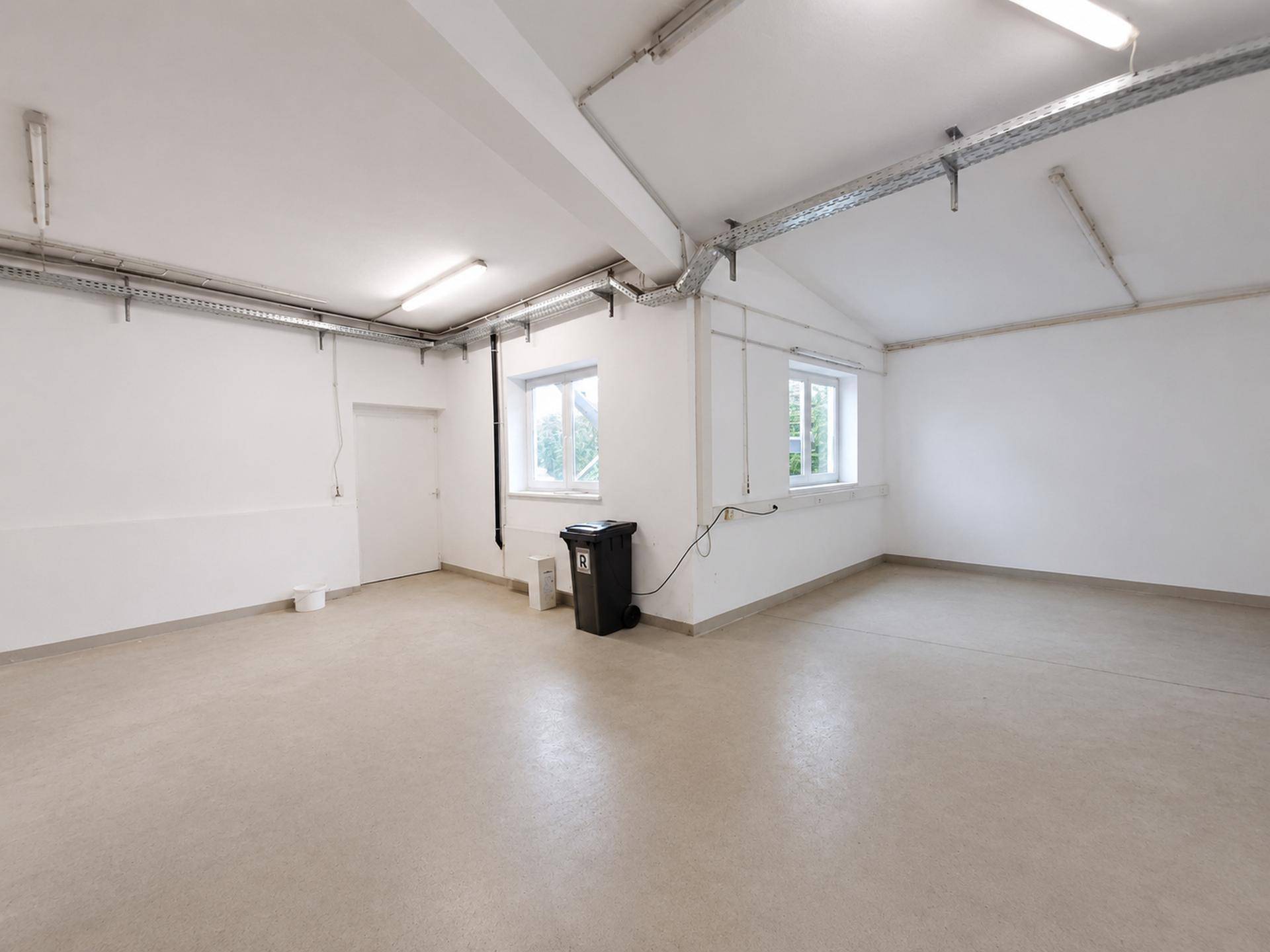 Heller Gewerberaum mit zwei Fenstern, Betonboden und sichtbaren Installationen an der Decke.