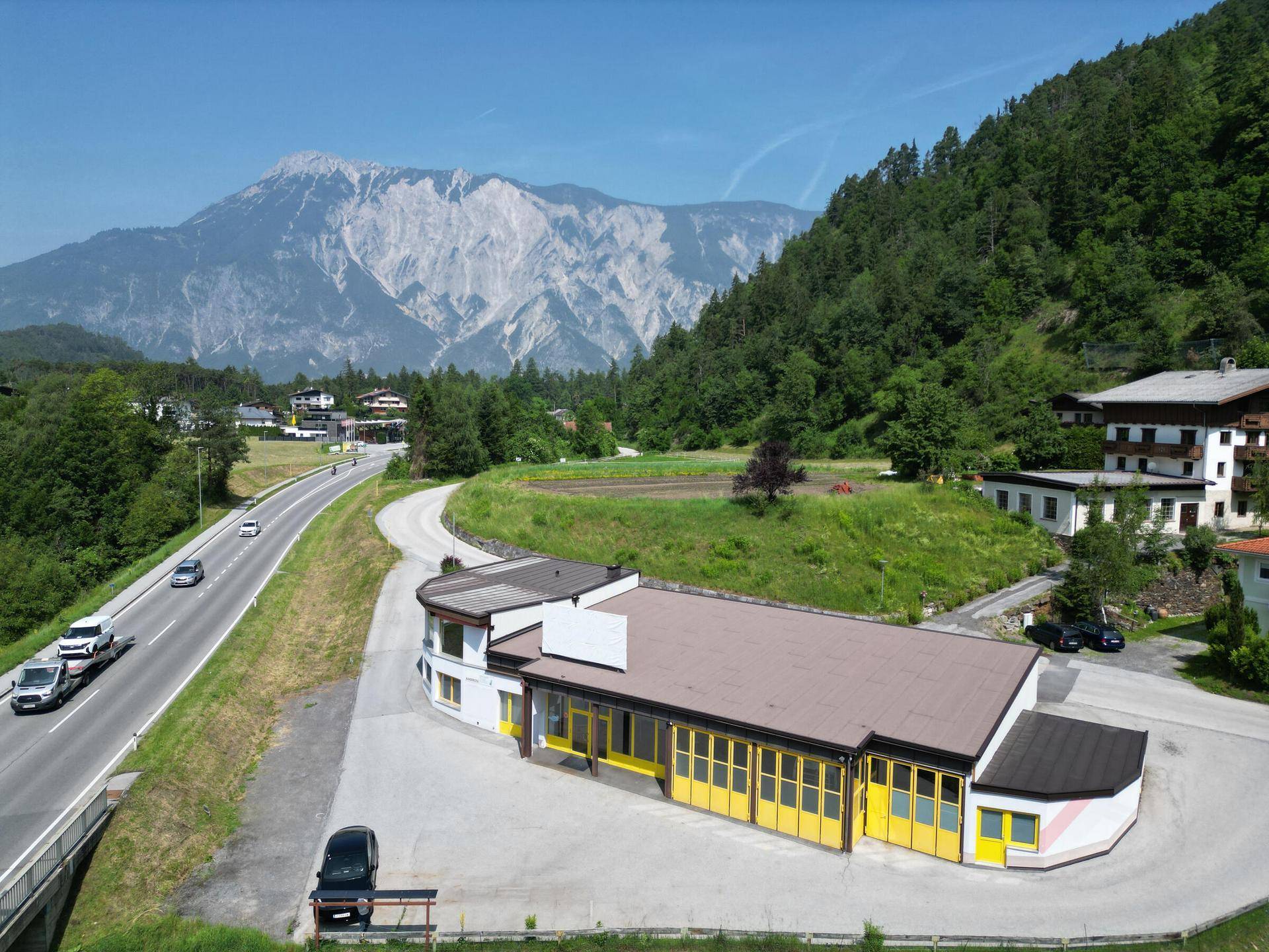 Luftaufnahme eines Gewerbegebäudes an einer Straße mit beeindruckender Bergkulisse und viel Grün im Hintergrund.