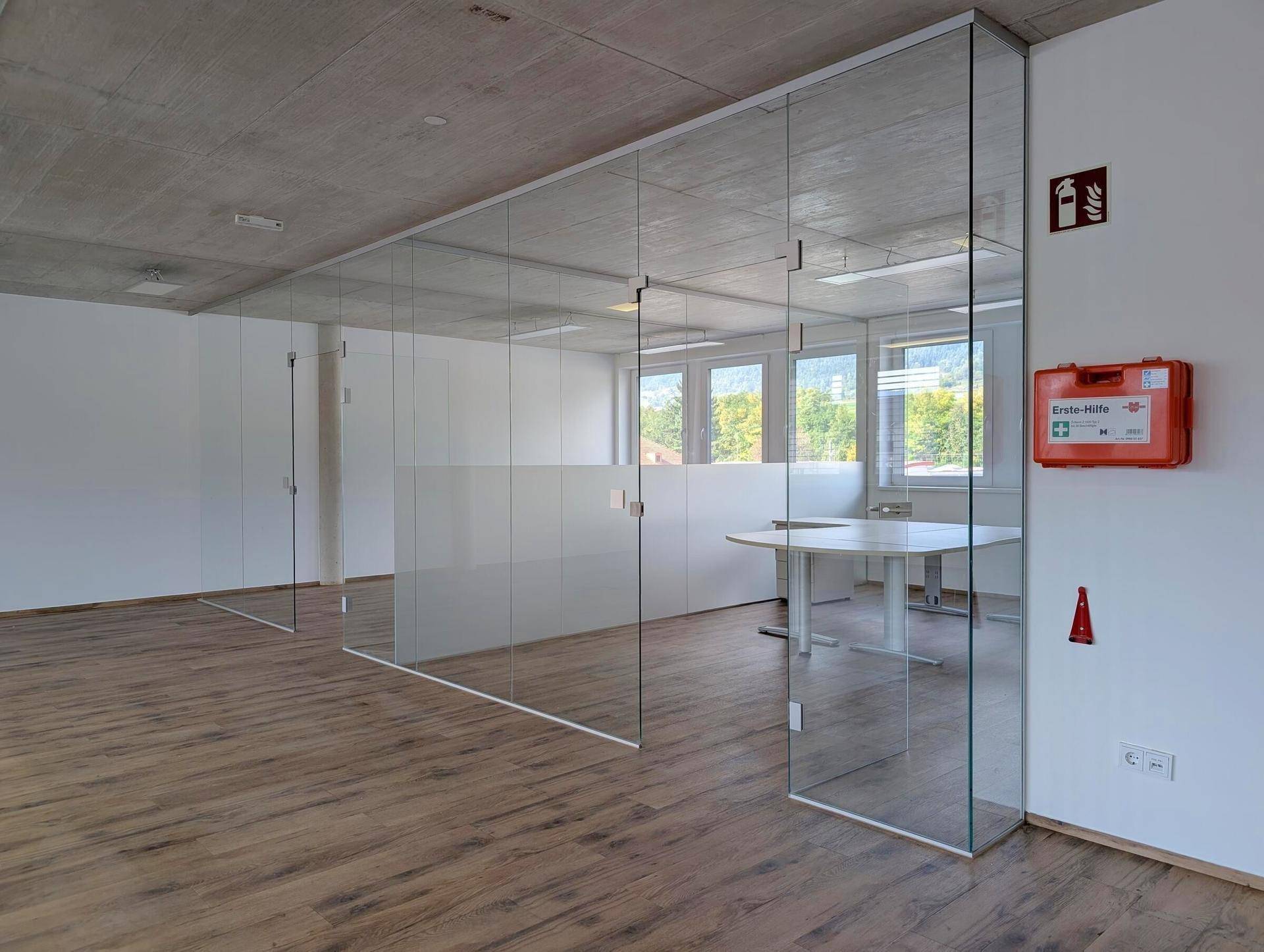 Modernes Büro mit Glasabtrennungen, Holzboden und viel Tageslicht, ausgestattet mit einem Erste-Hilfe-Kasten.