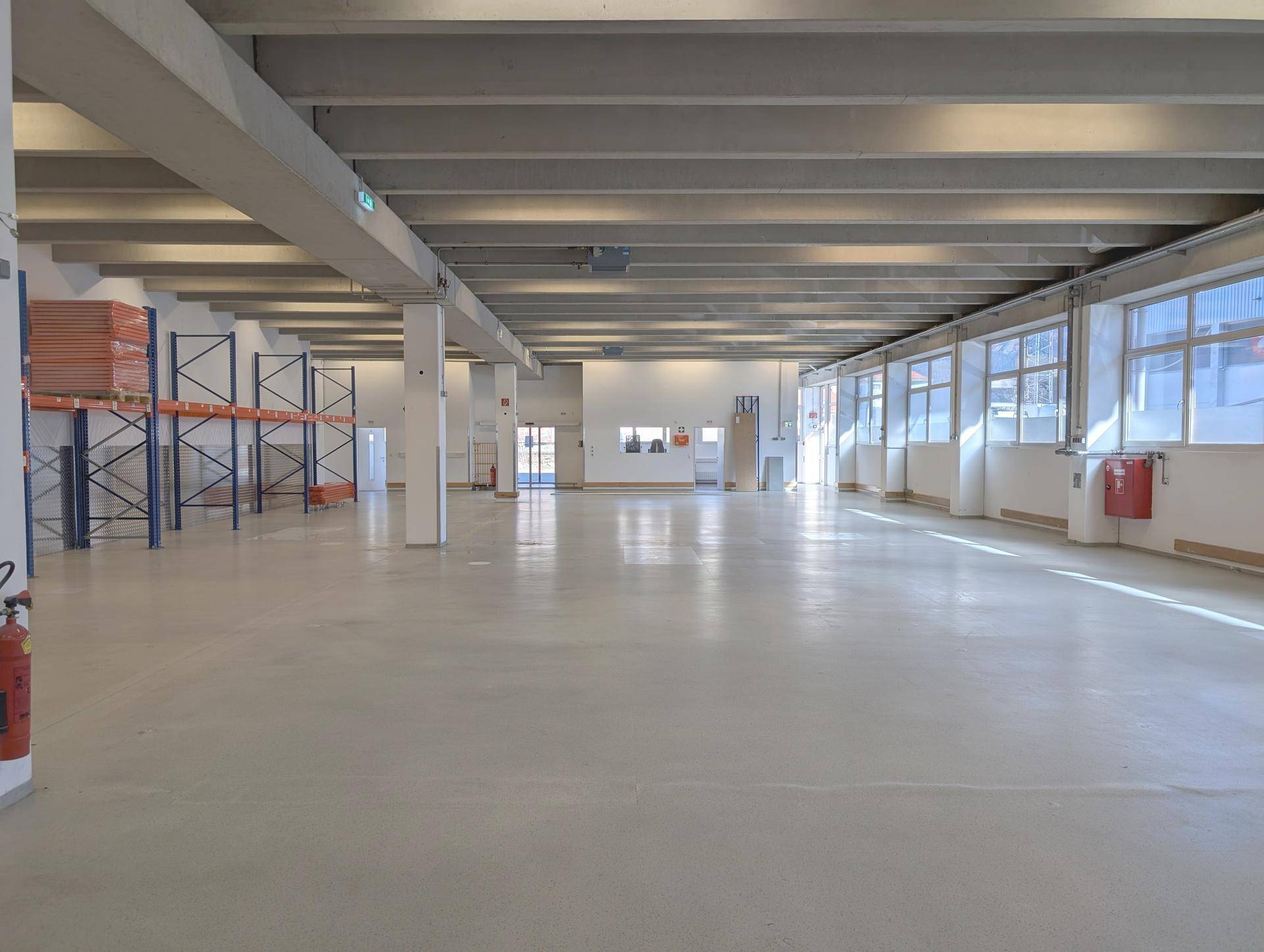Geräumige, leere Lagerhalle mit hohen Decken, Betonboden und natürlichem Lichteinfall durch Fenster.