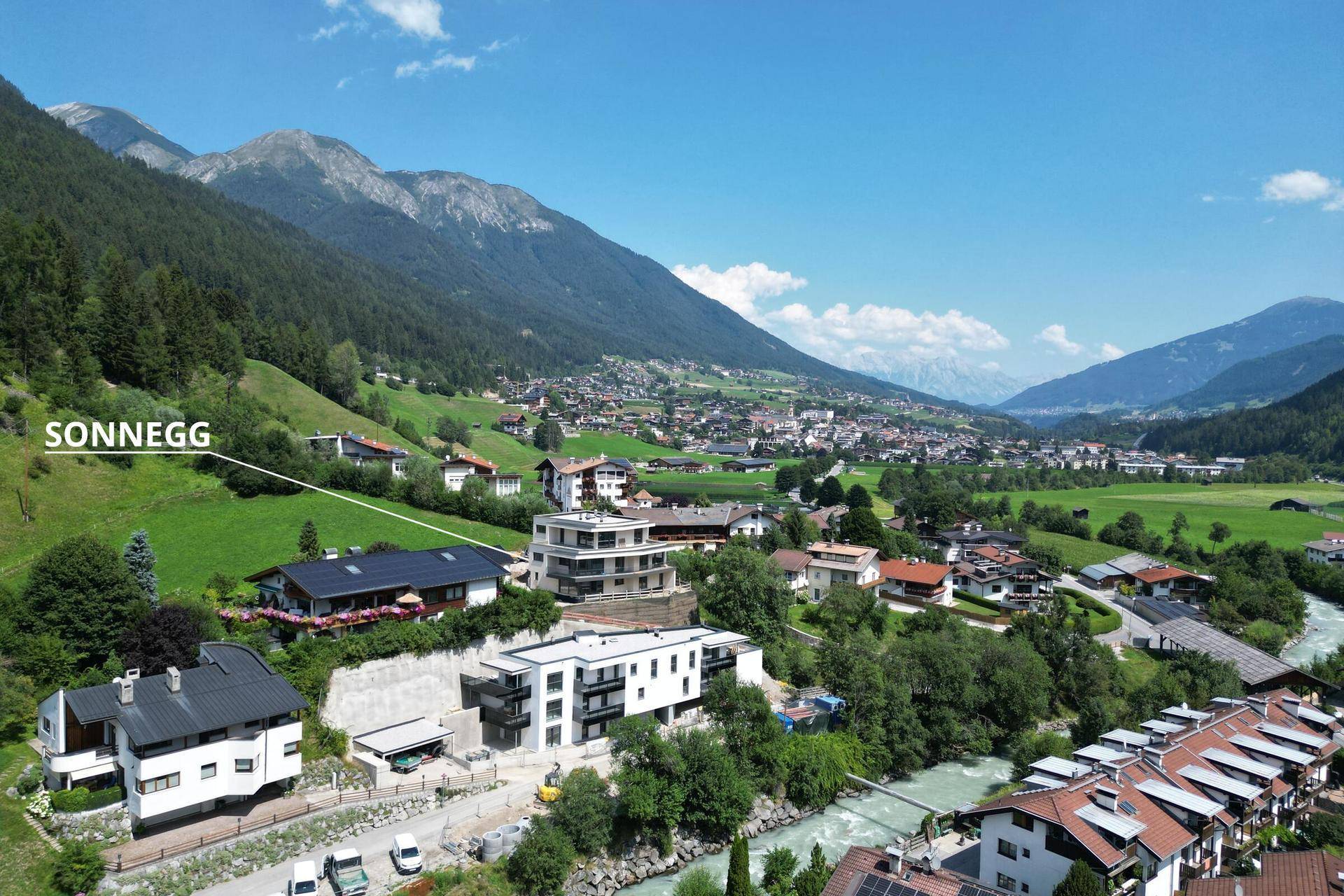 Weitläufige Luftaufnahme eines Neubaus in einem grünen Tal, umgeben von Bergen und einem Fluss.