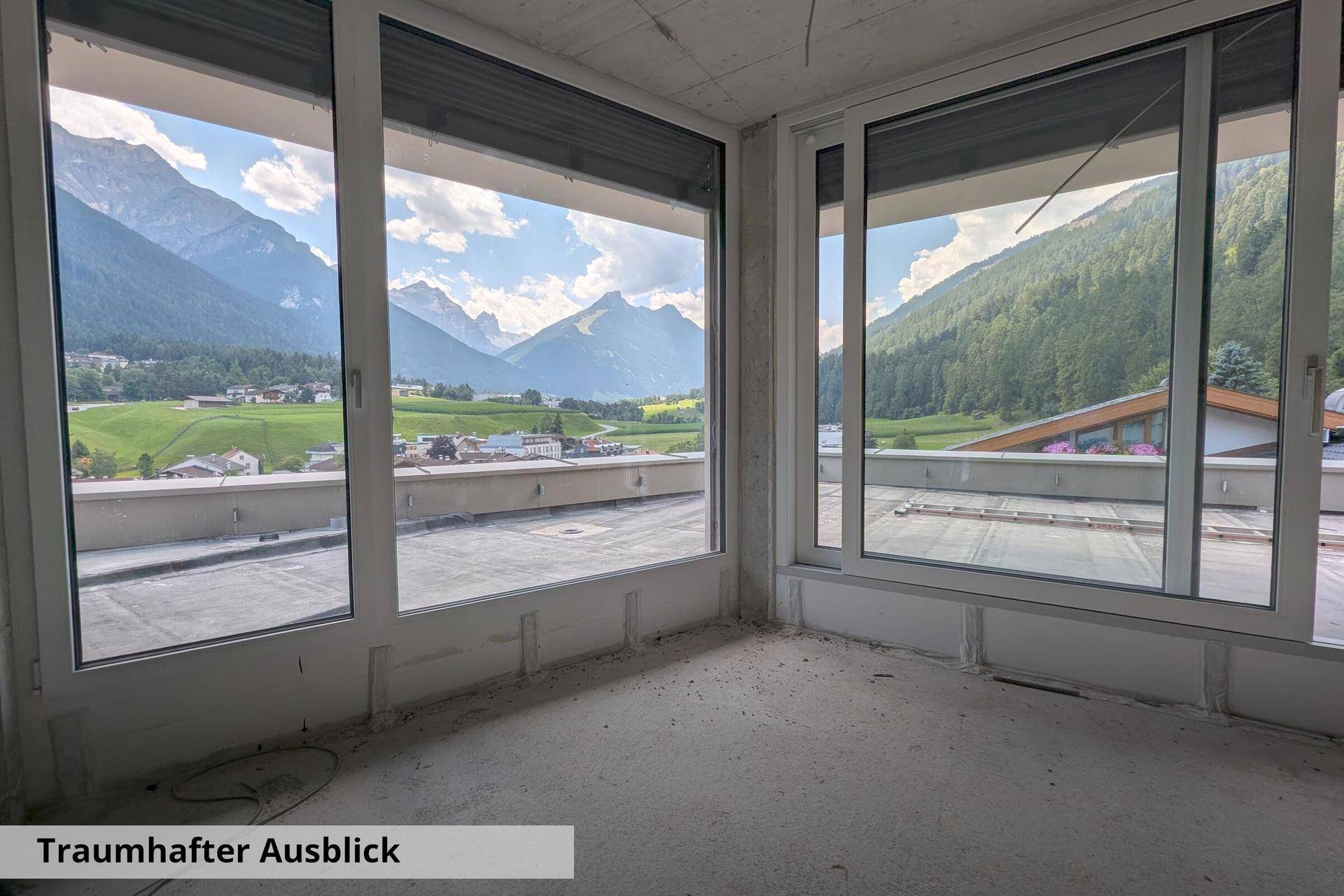 Innenraum im Rohbau mit großen Fenstern, die einen weiten Blick auf die alpine Landschaft bieten.