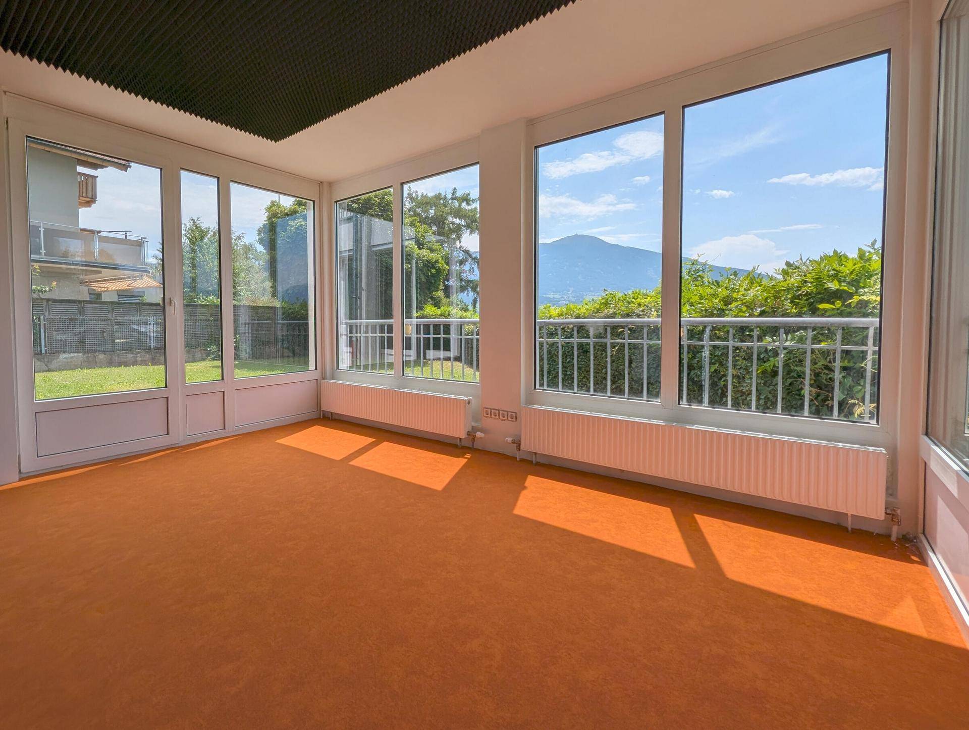 Heller Innenraum mit weitem Bergblick durch große Fenster, ausgestattet mit orangefarbenem Teppich und Akustikdeckenpaneelen.